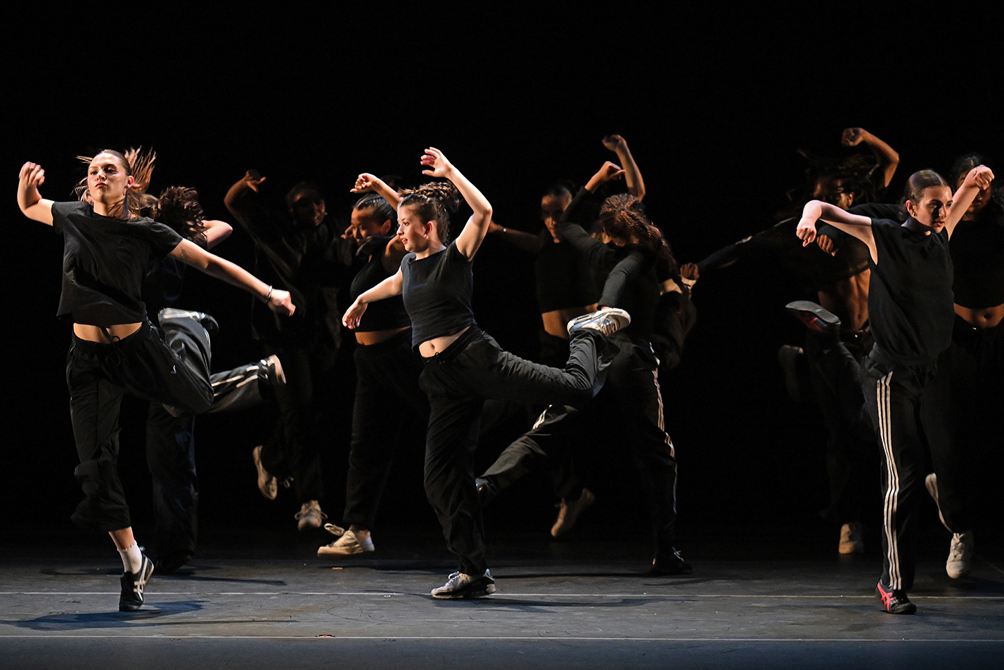 Modern dancers in black pants and black tank tops perform in a group with high arm motions and high steps.