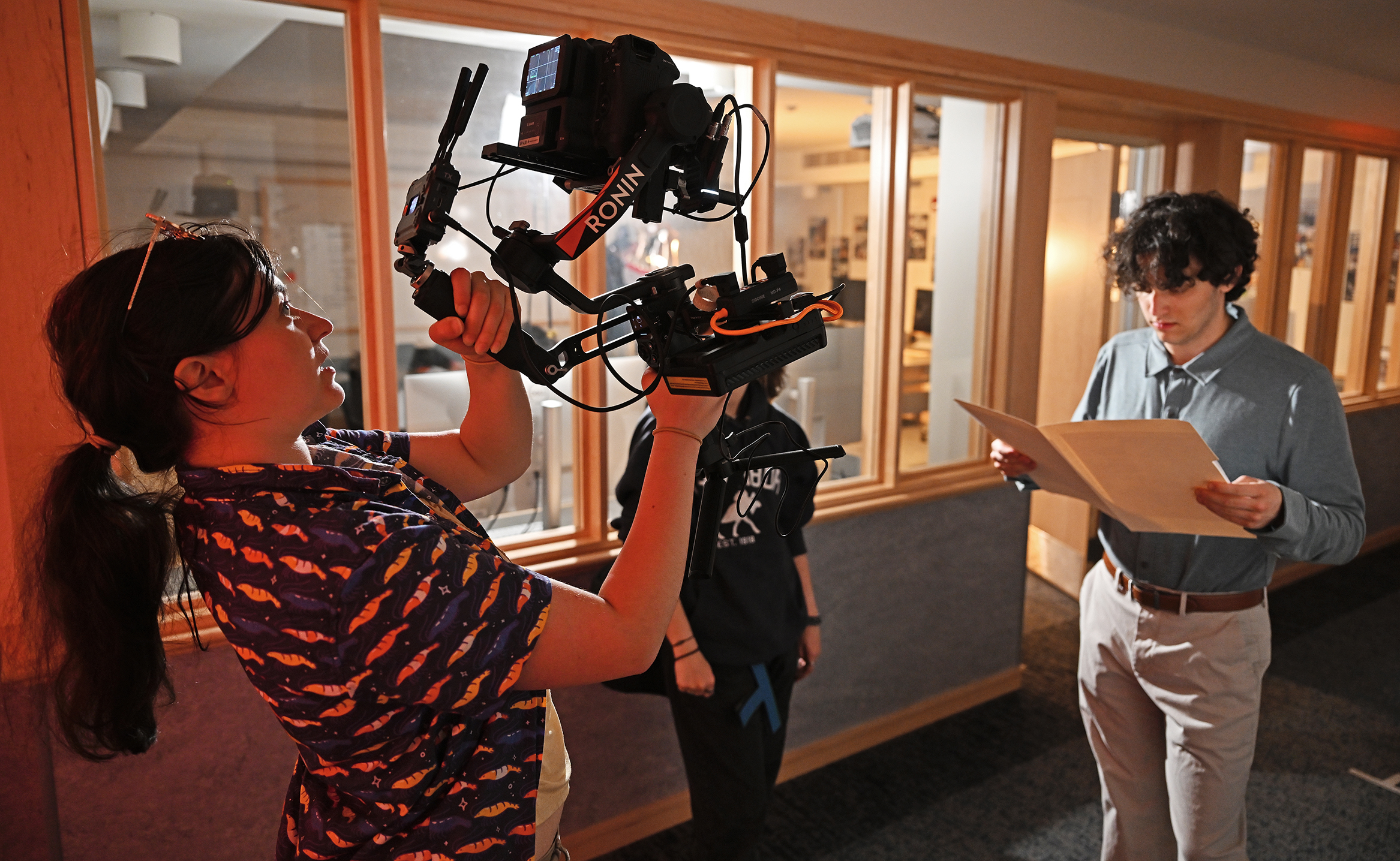 A female film student with pony tail and glasses atop her head holds a large video camera perched on a gimbal as an actor looks a papers in a manila folder in a dimly lit hallway in front of a row of windows during a film class.