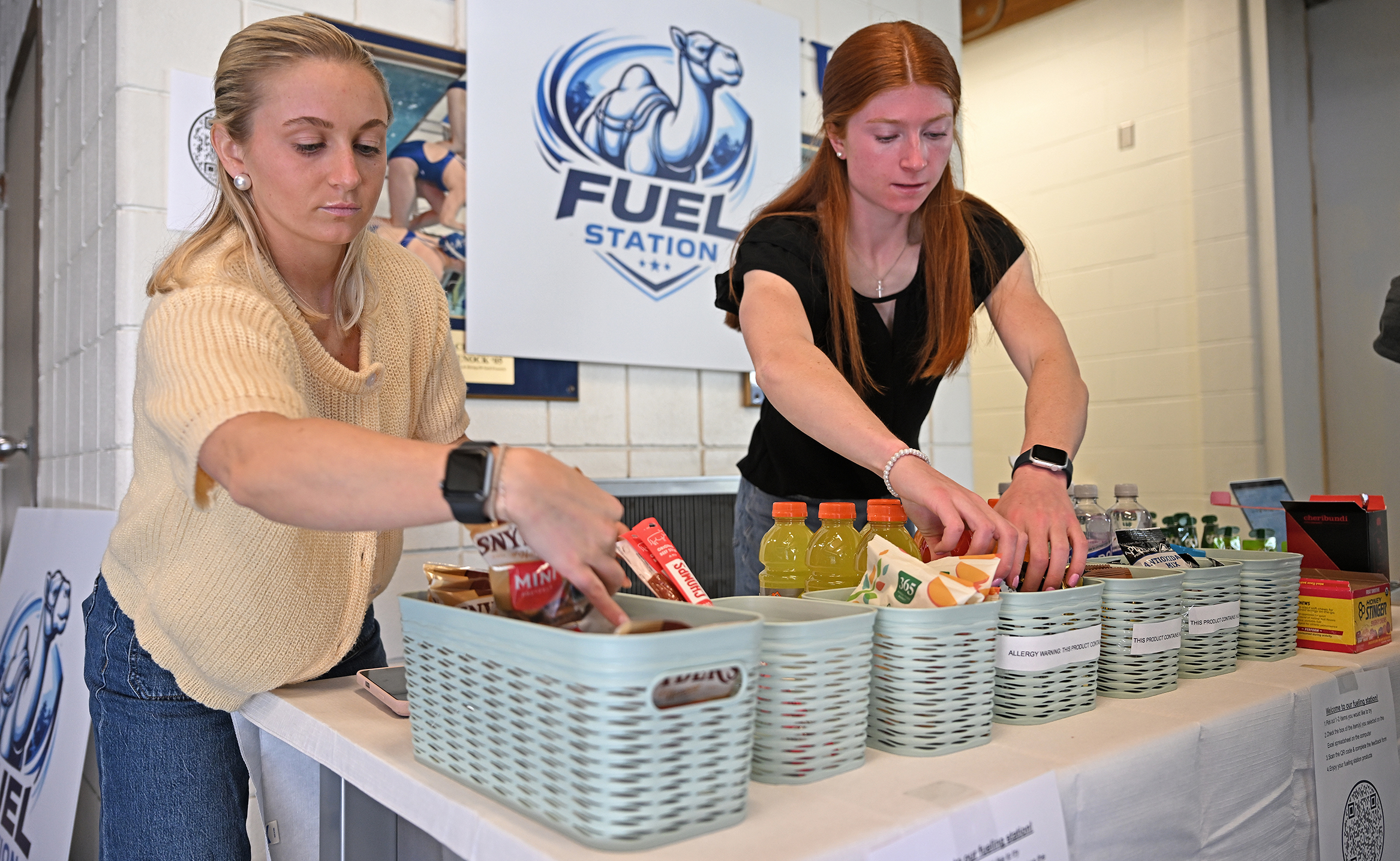 Two female students, one blond, one redhead, add snacks to baskets on a table in front of a sign showing a camel and the words 