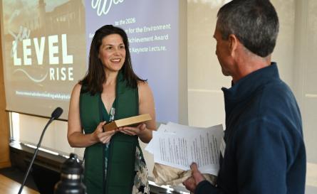 A female speaker wearing a sleeveless top and a wide green silk stole holds an award plaque presented to her by a male professor holding a sheaf of papers in front of a projection screen with the words 