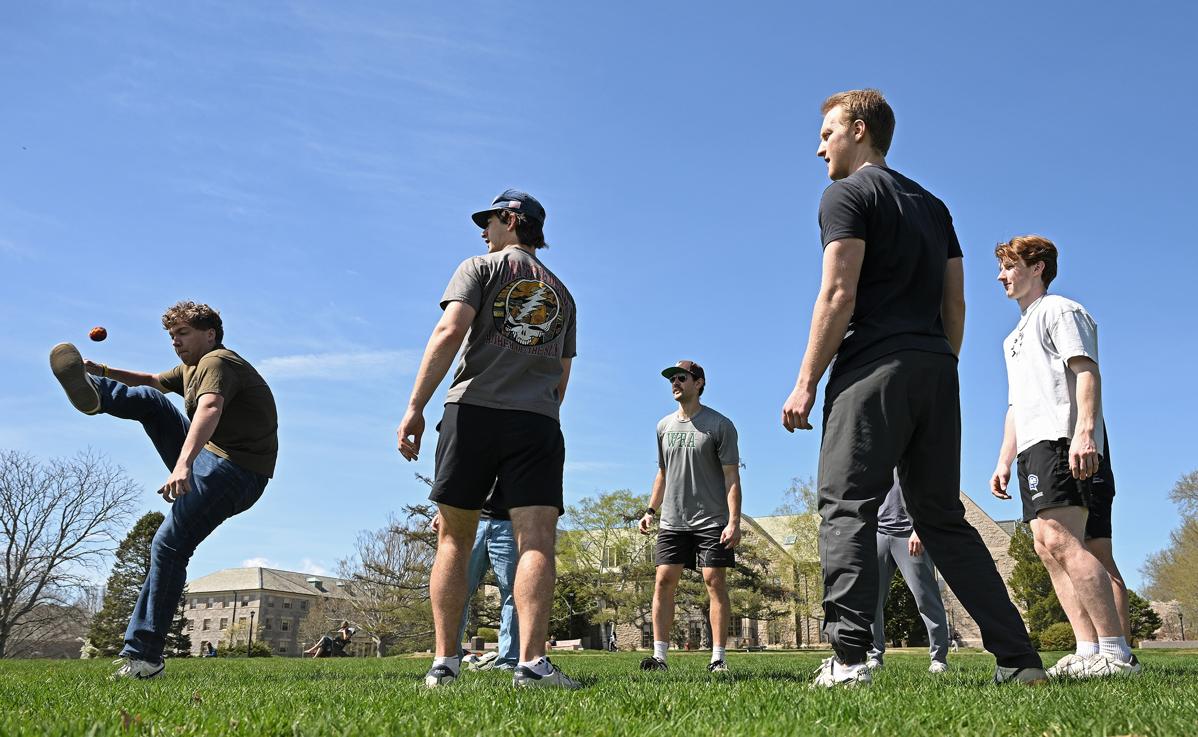A group of male students, with one on left kicking a foot high in the air, play hackey sack on a verdant college green with granite buildings visible in the distance.