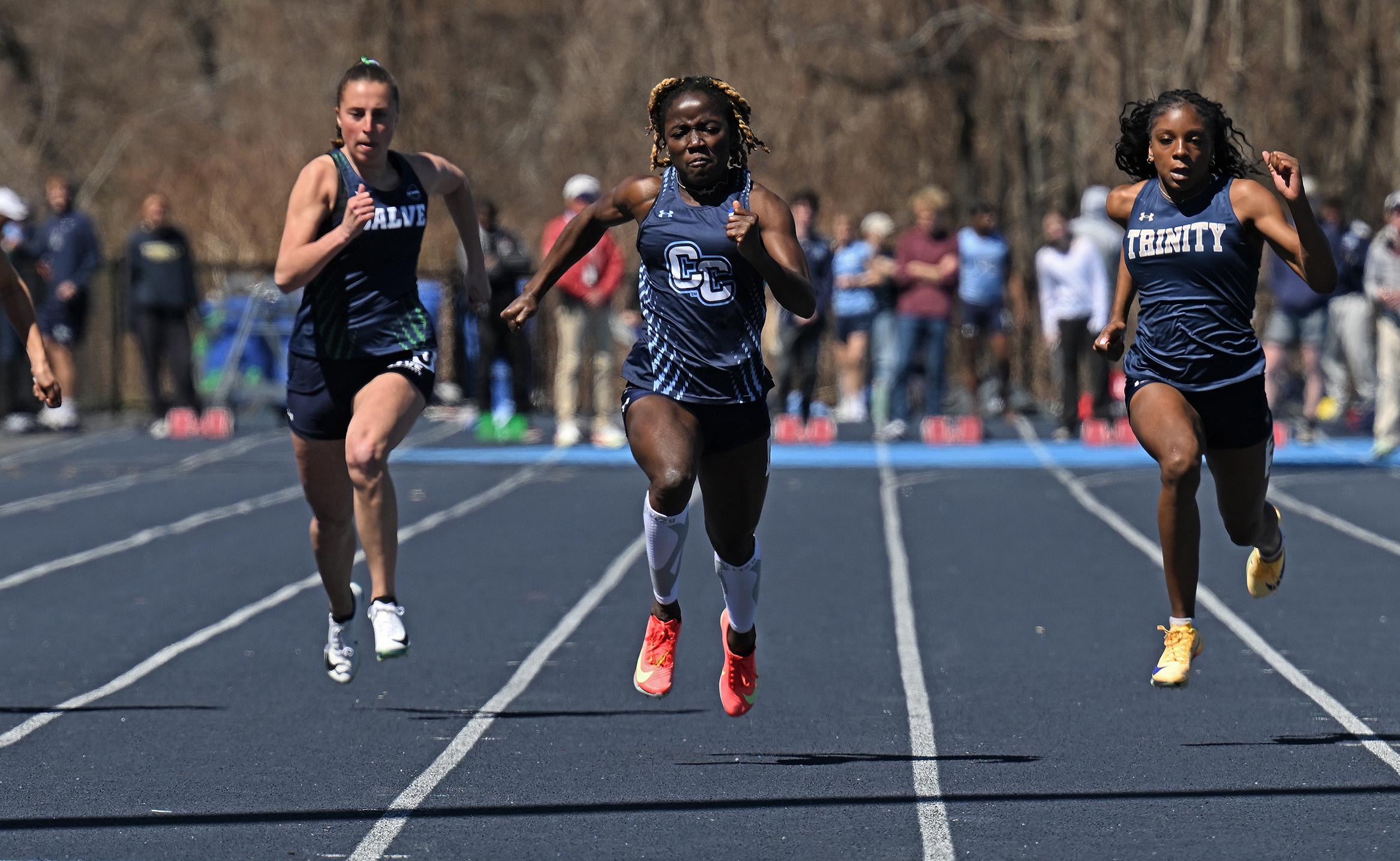 Three female college athletes spring toward the finish line in a 100 meter race on a blue track.