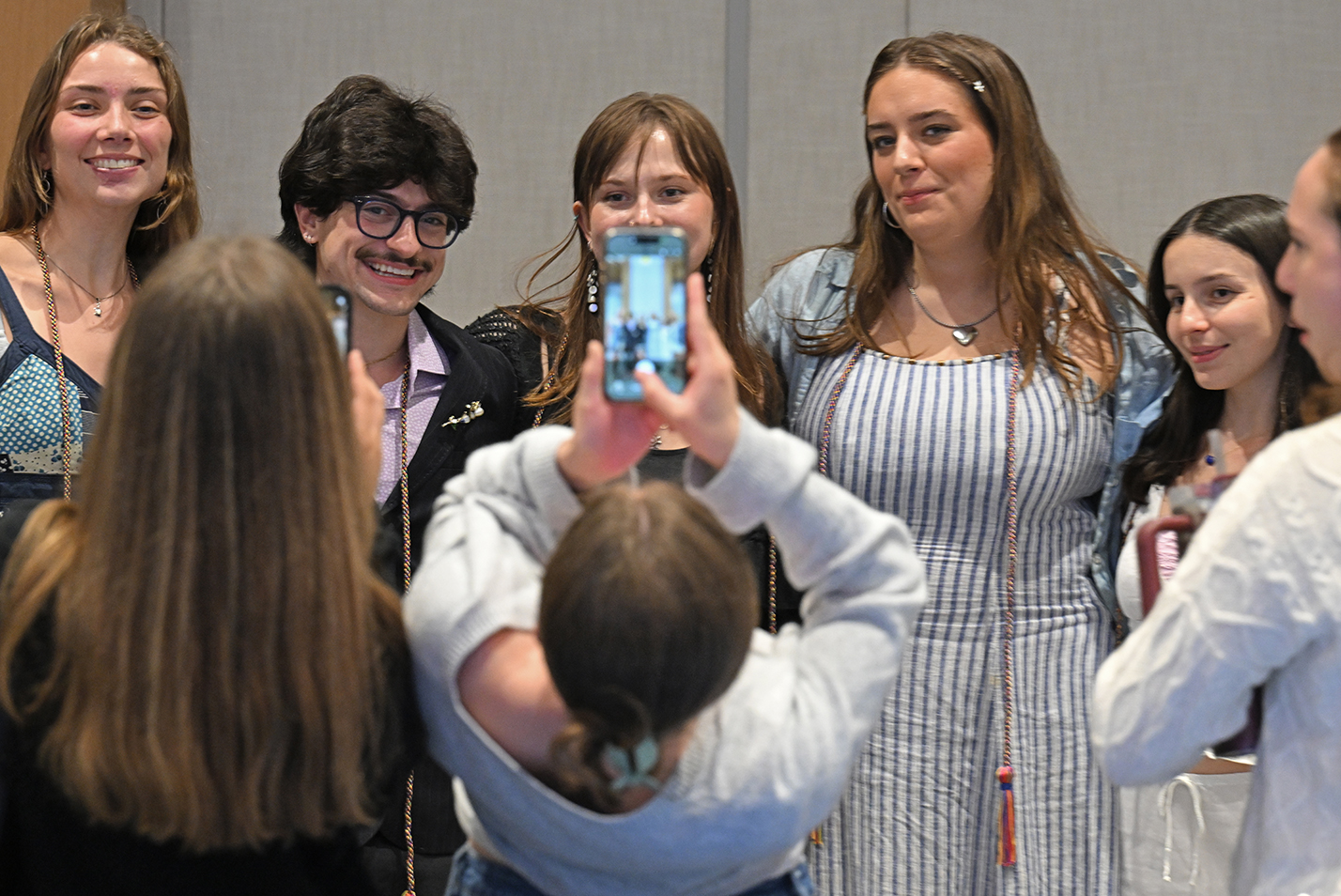 A group of students sporting rainbow graduation cords pose for a photographer who is leaving back to fit them all in the screen.