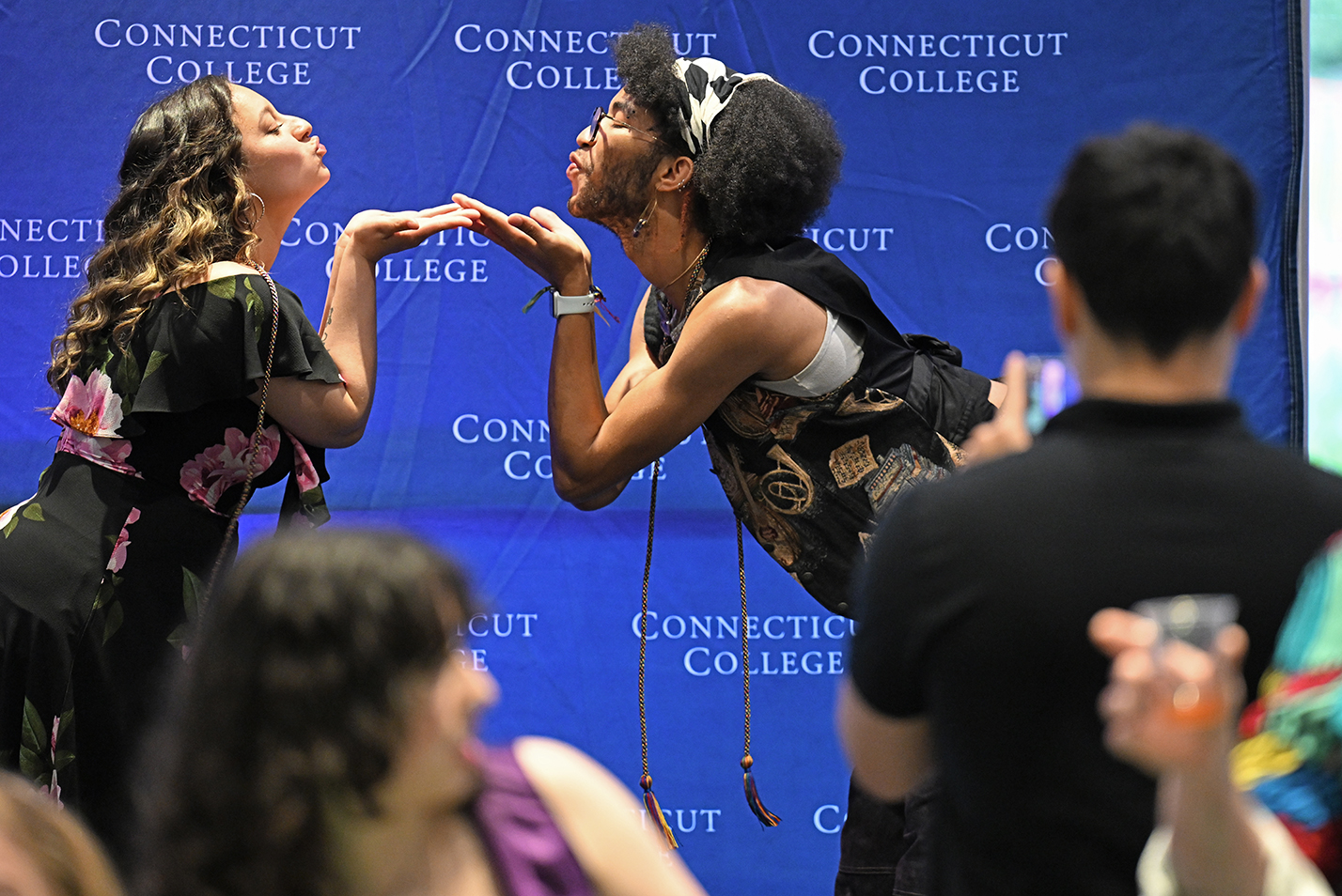 Two students lean toward each other pantomiming blowing kisses as they pose for a classmate with a smart phone camera.