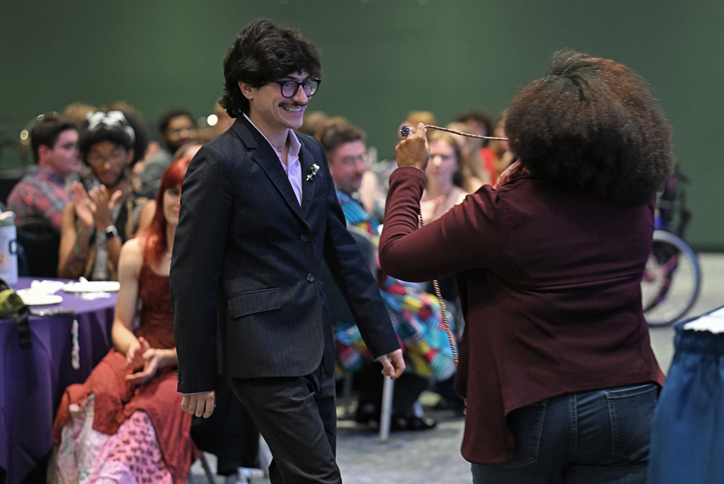 A male student with a mustache and neatly coifed hair steps out of the audience to approach a college administrator presenting a rainbow graduation cord.