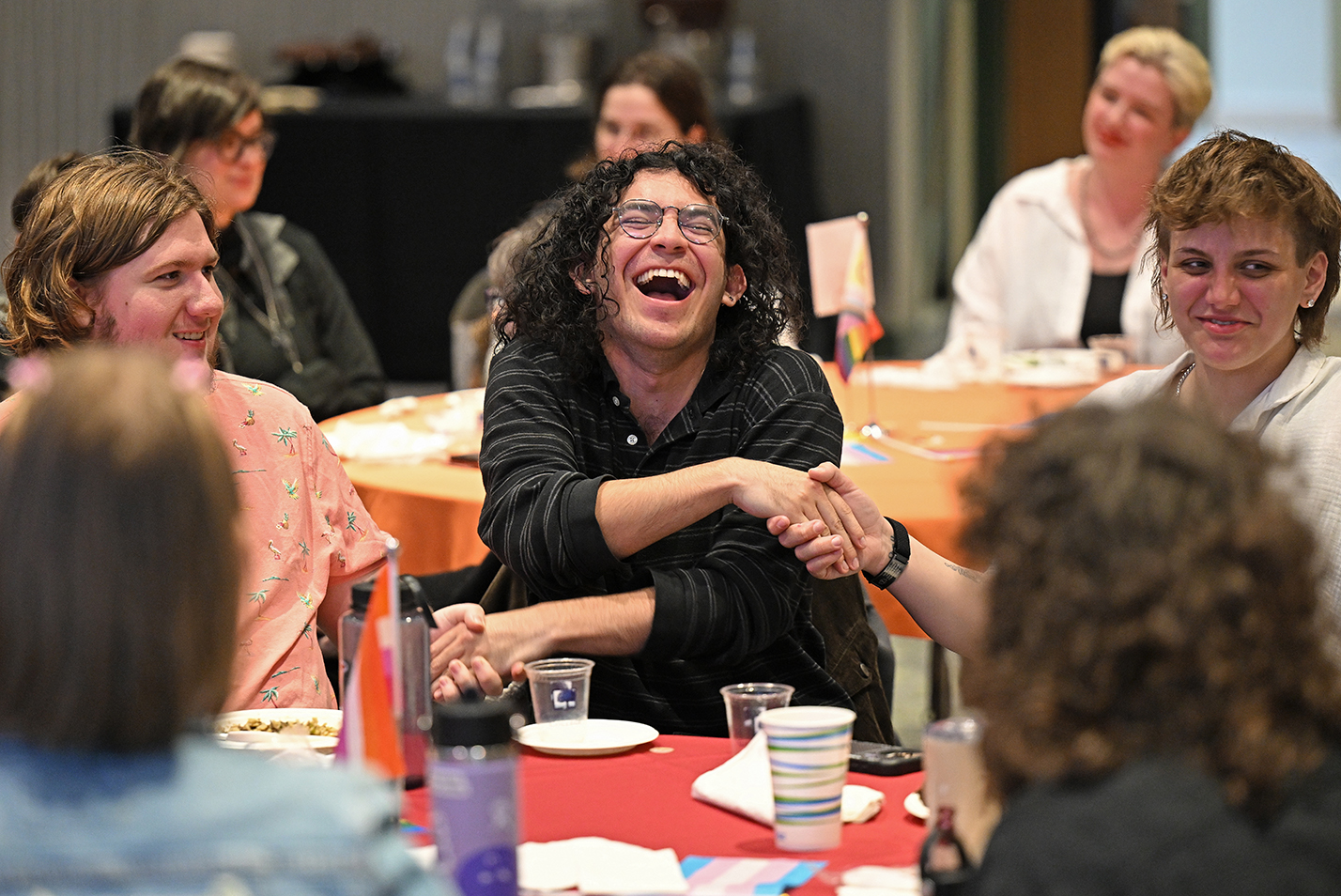 A student with long curly hair laughs as he crosses his arms to hold hands with people on either side while seated at a table.
