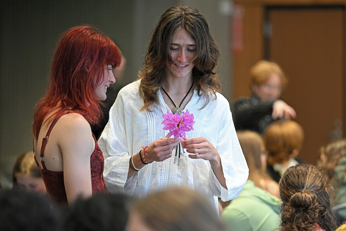 Two students with long hair, one holding a bright pink flower, stand amidst a seated crowd talking quietly to each other.