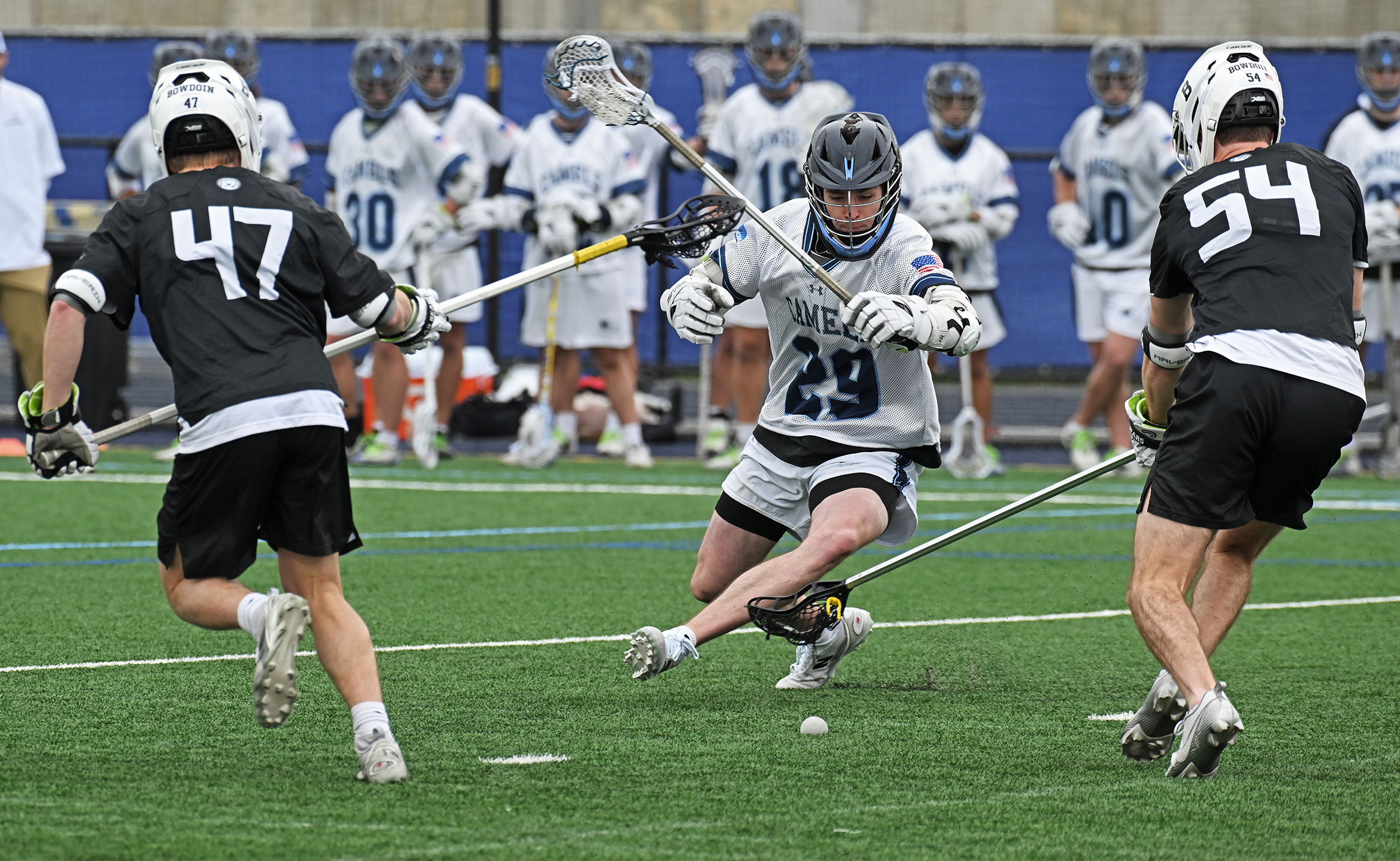 A male lacrosse player in white swings his stick one handed at a ball on the ground as two players in black also reach for the ball.