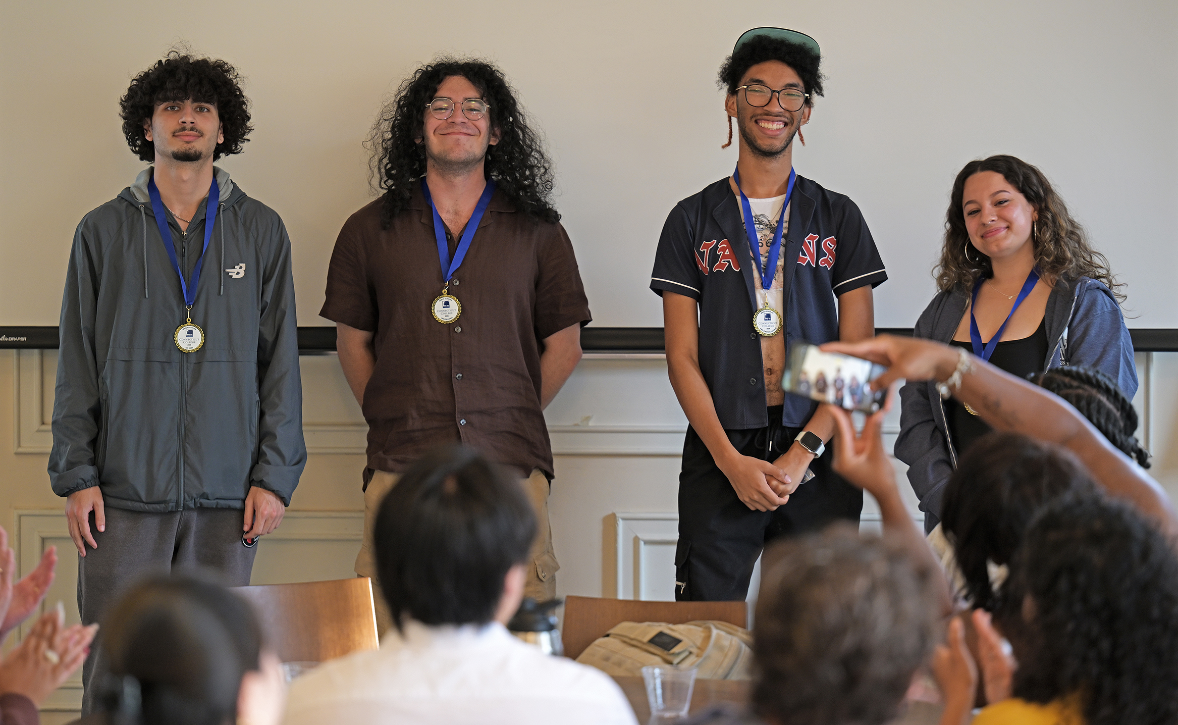 Four college students stand wearing medals as a member of the audience holds up a smart phone to take their photo.