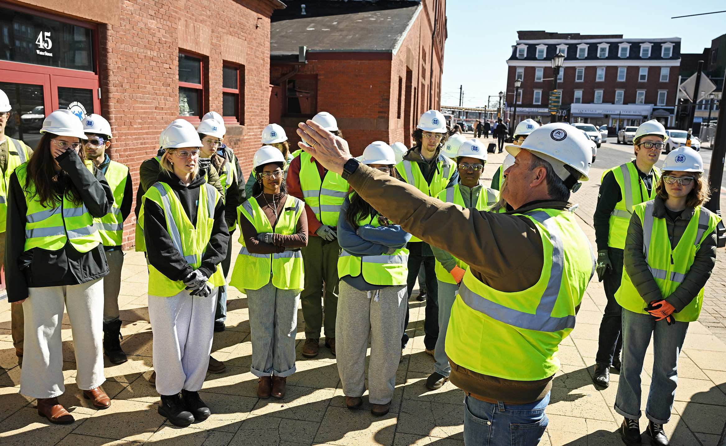 A man in a white hardhad and yellow safety vest gestures with his left hand speaking to a group of college students, also in white hardhats and yellow safety vests, while standing outside a brick building.