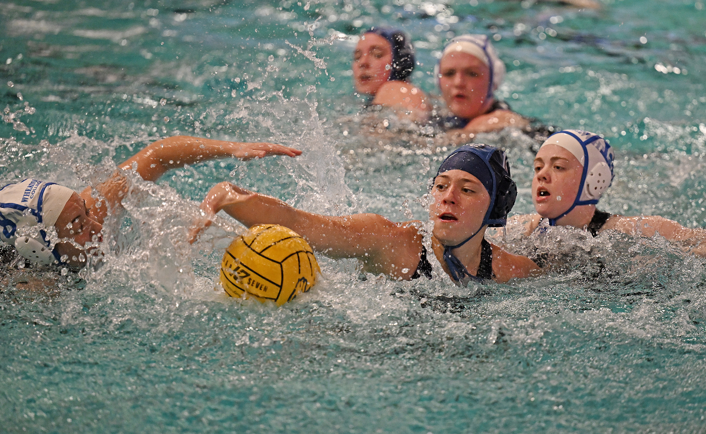 A female water polo player in blue reaches for the ball as opposing players in white surround her.