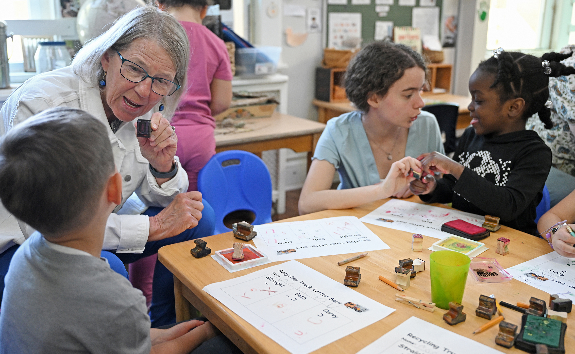 An older woman with mid-length greying hair holds a letter stamp talking to a preschool student as a college student does the same thing in the background.