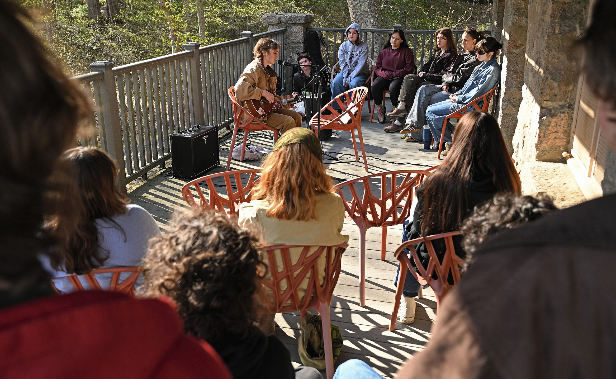 A student performing on acoustic guitar sings for an audience seated around them on a deck surrounded by woods.