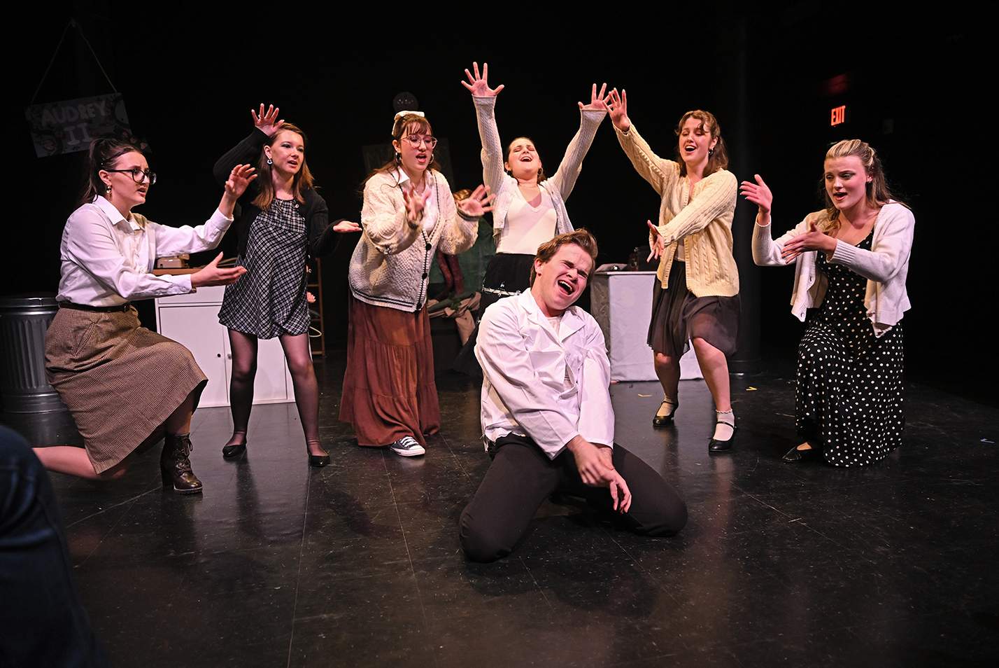 An actor in a white lab coat kneels on the floor singing while surrounded by the ensemble all gesturing with their arms.