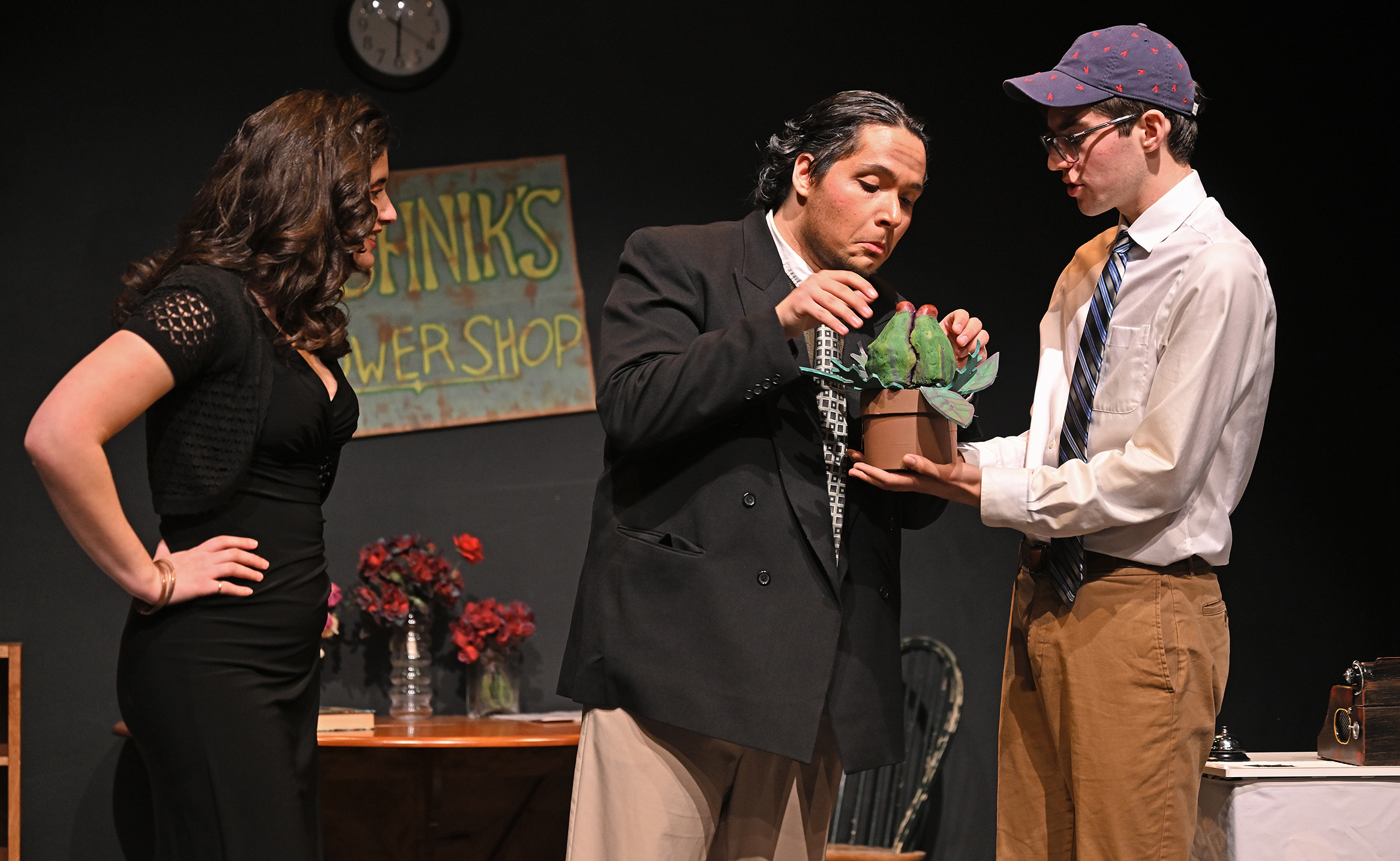 A student actor with slicked back hair and suit jacket makes a face while examining a prop plant that looks like a comical Venus Flytrap help by the actor in white shirt and baseball cap, as another actor in a tight black dress looks on.