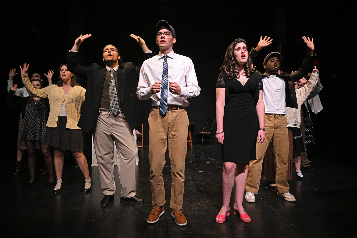 The cast of a musical production perform a group song standing in a semi-circle on the stage in a black box theater.