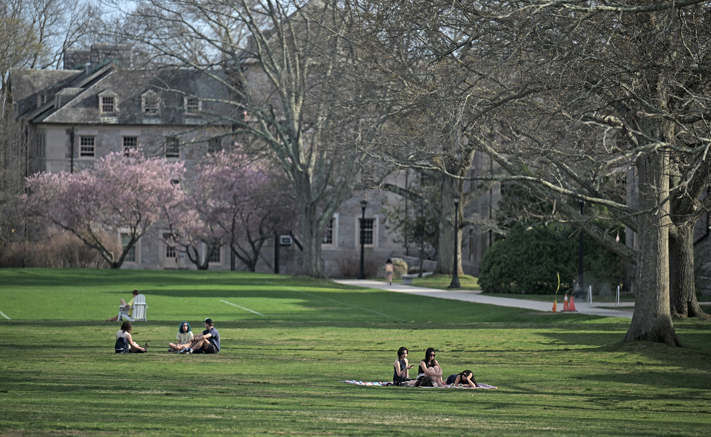 College students relax on blankets and Adirondack chairs scattered about a verdant college green on a mild sunny afternoon.
