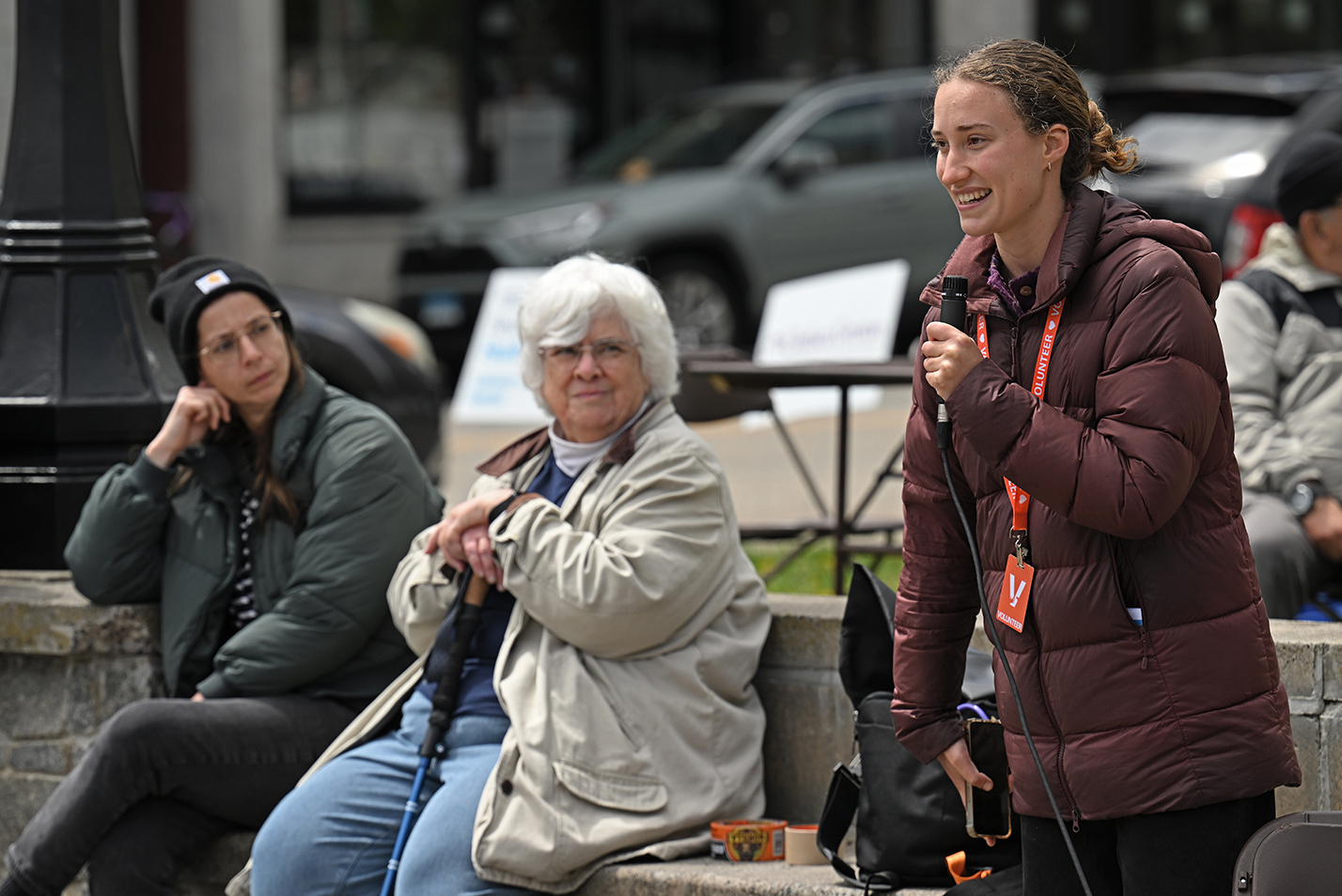 A student in a brown winter coat holds a microphone and addresses a crown as a kindly woman with short white hair watches from the background.