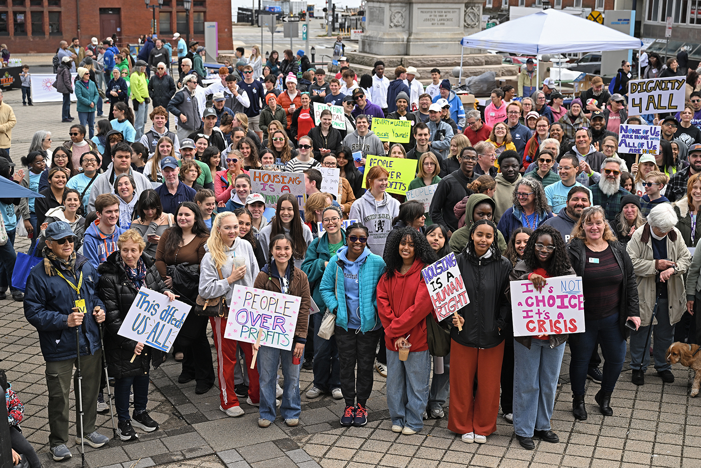 A large group of people for an end homelessness march gather in a city plaza holding signs.