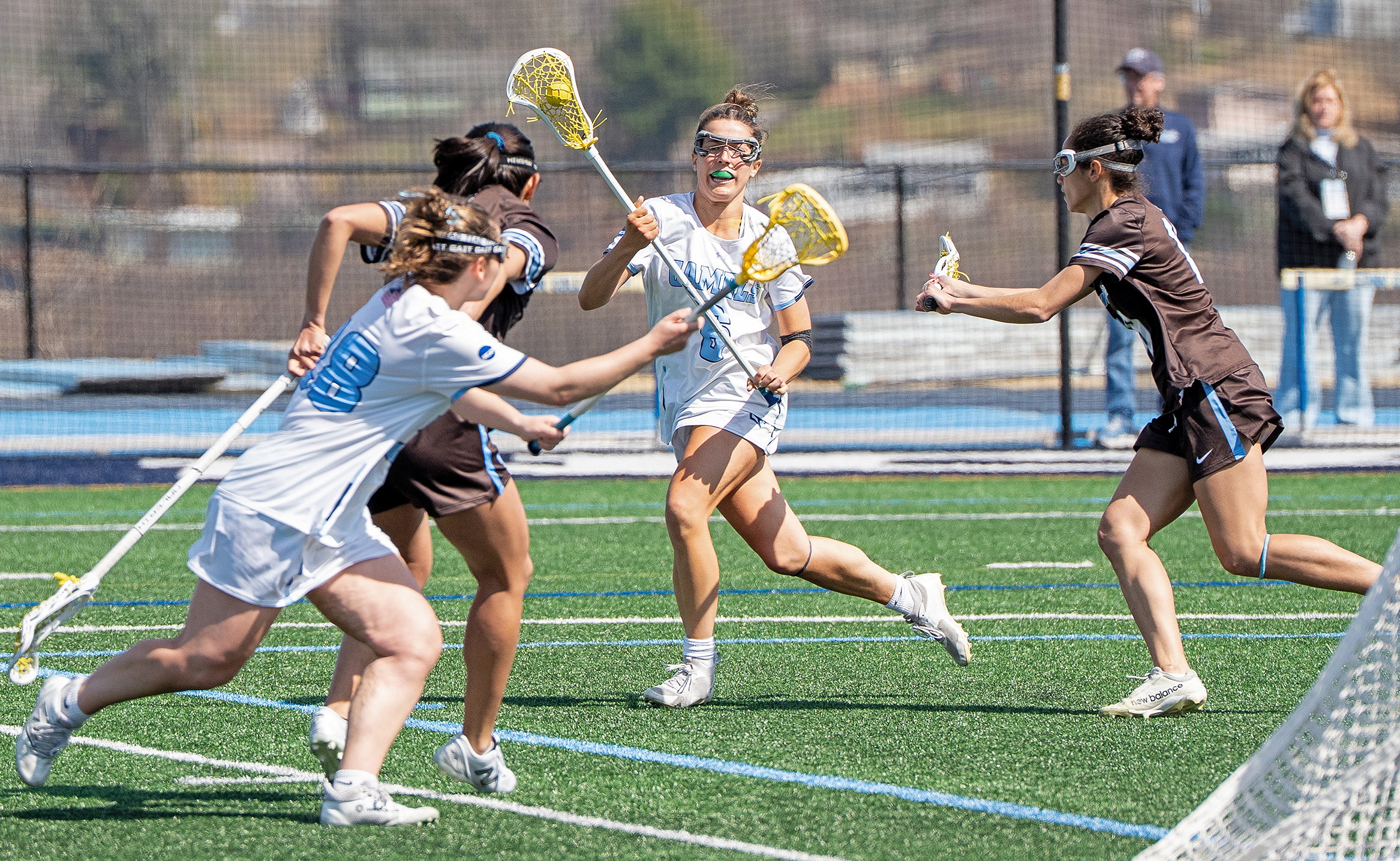 A women's lacrosse player at full stride passes the ball overhand to a teammate in the foreground cutting toward the goal with an opponent in pursuit.