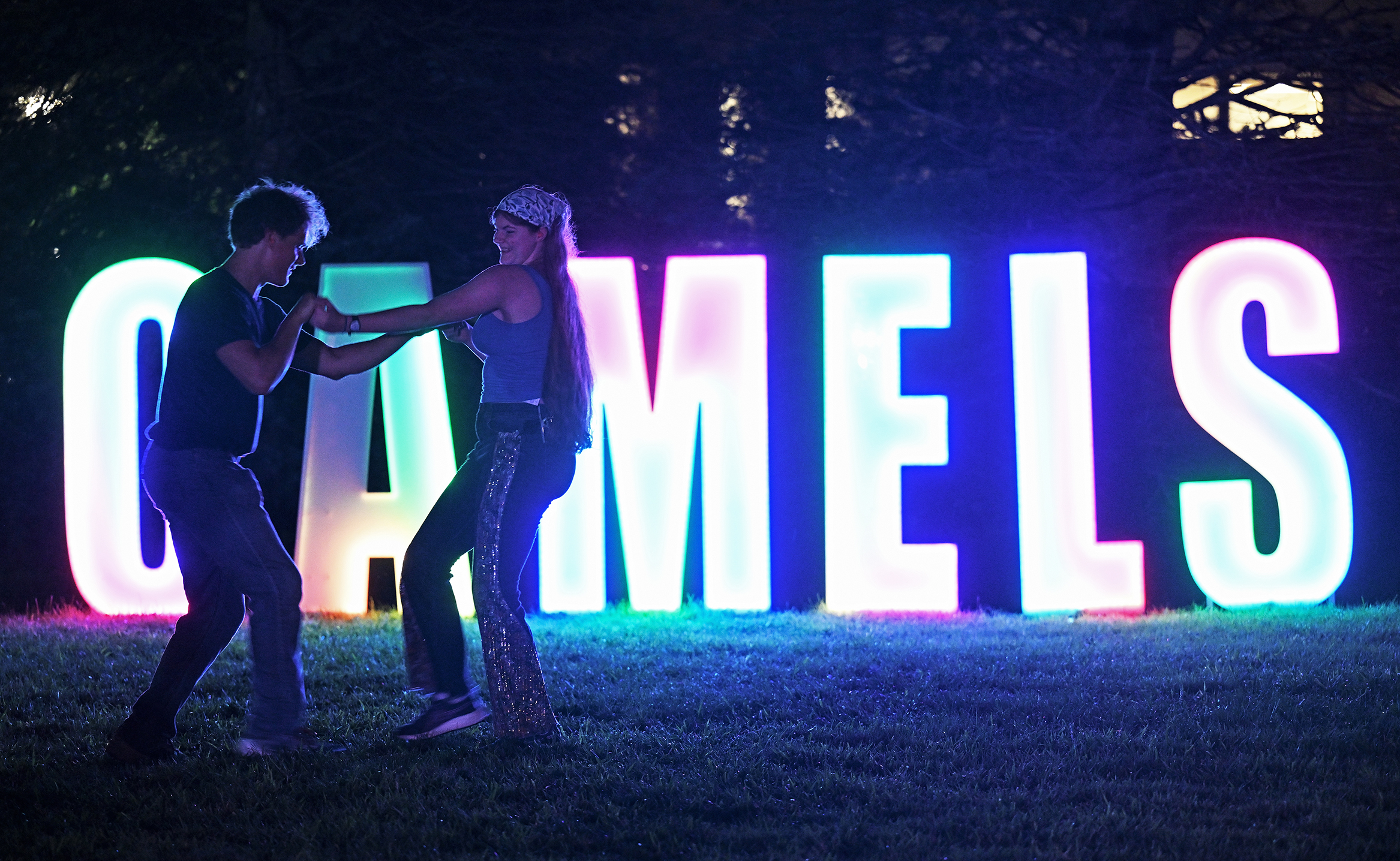 Two college students dance after dark on a campus green in front of giant lighted letters spelling out CAMELS.
