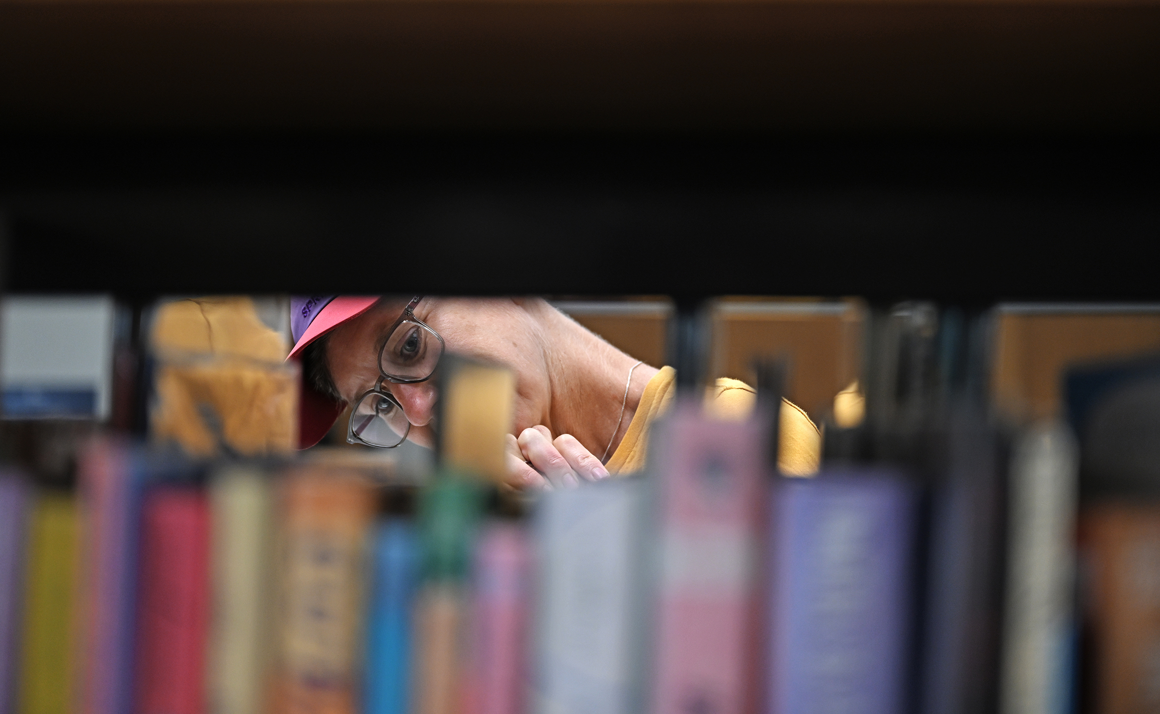 A college professor with a pink visored ball cap and glasses can be seen, head turned sideways, looking at books on a library shelf.