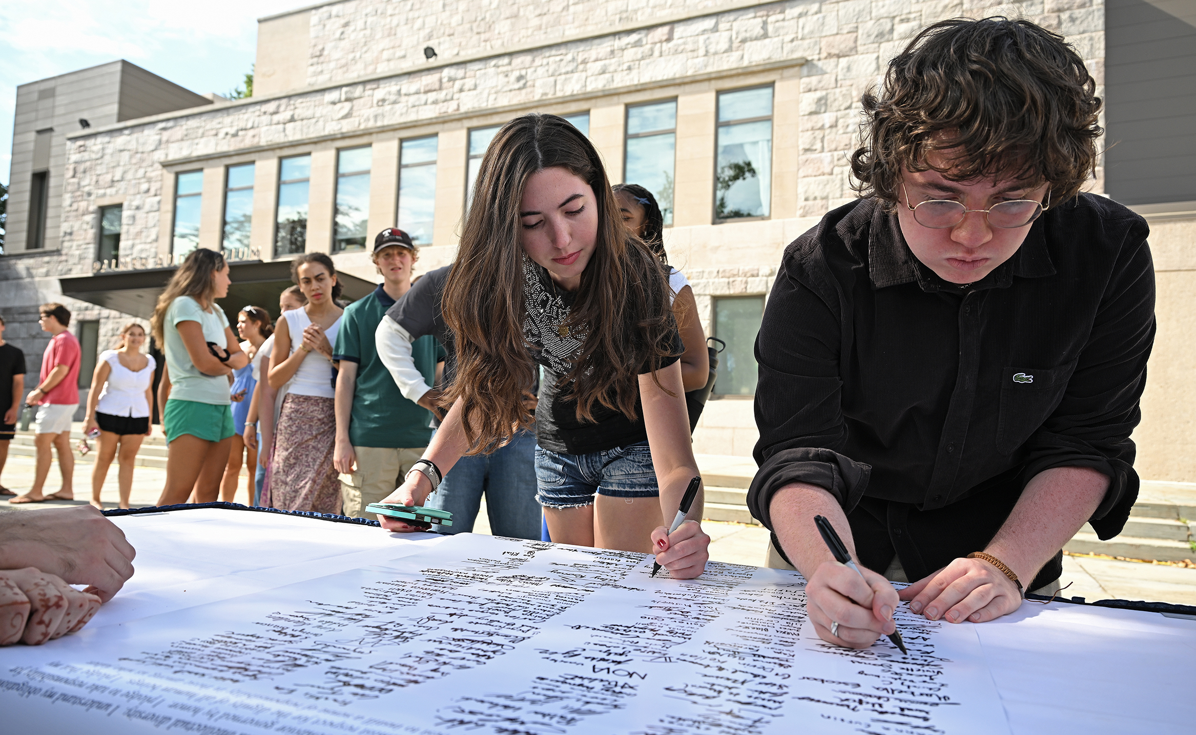 College students lean over to sign a poster-size document in an outdoor courtyard.