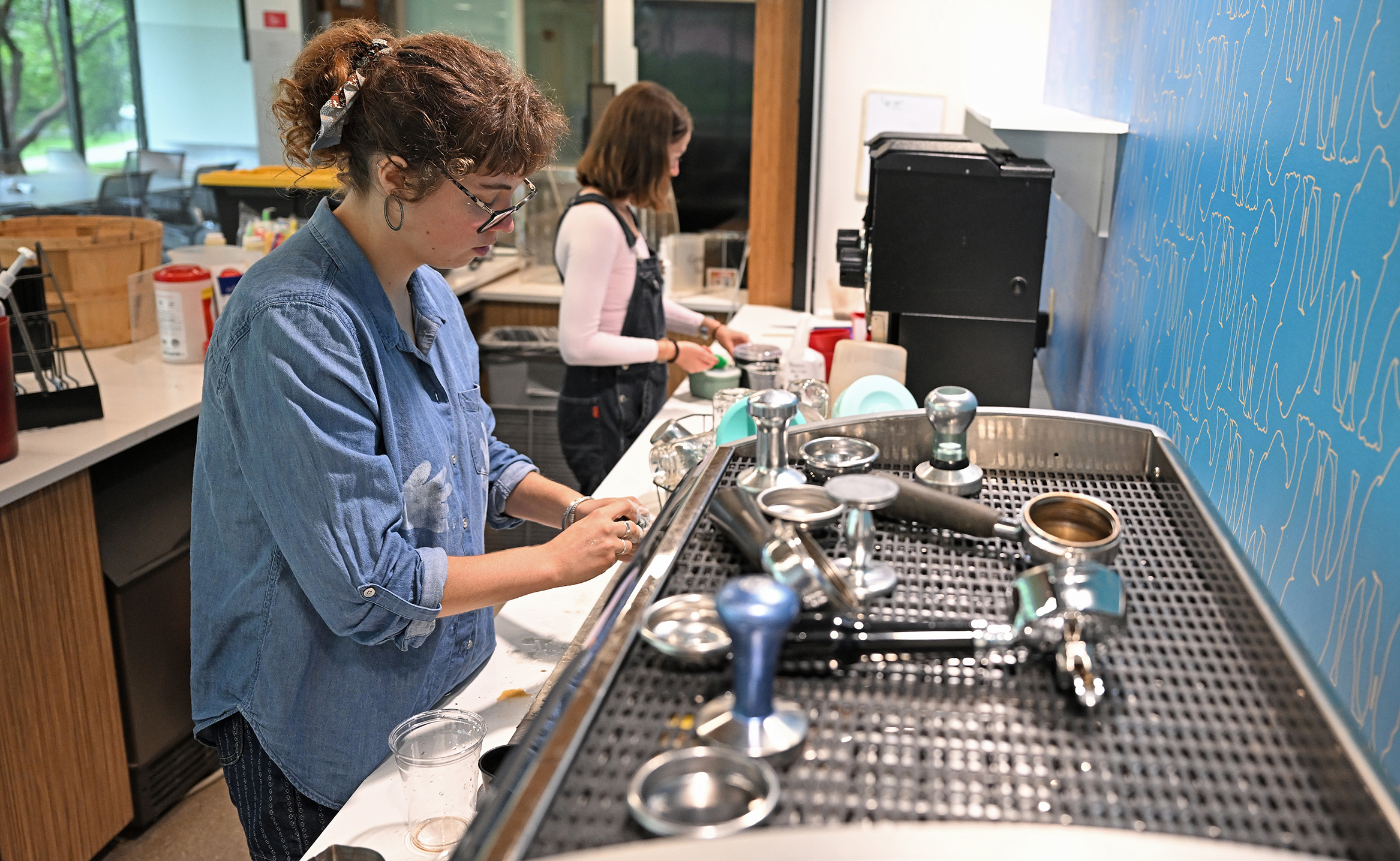 A college student coffee shop worker in denim shirt and glasses cleans the components of an espresso machine while another worker in overalls and pink shirt cleans the counter in the background.