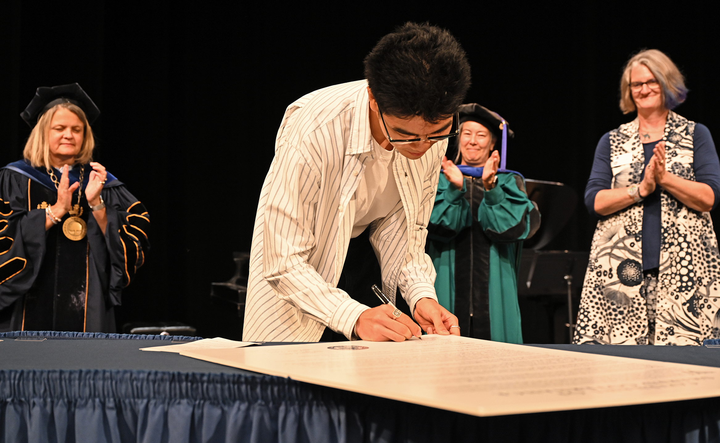 A college student in long sleep shirt leans over to sign a poster-size document as college personnel in academic regalia watch from the background on a stage.