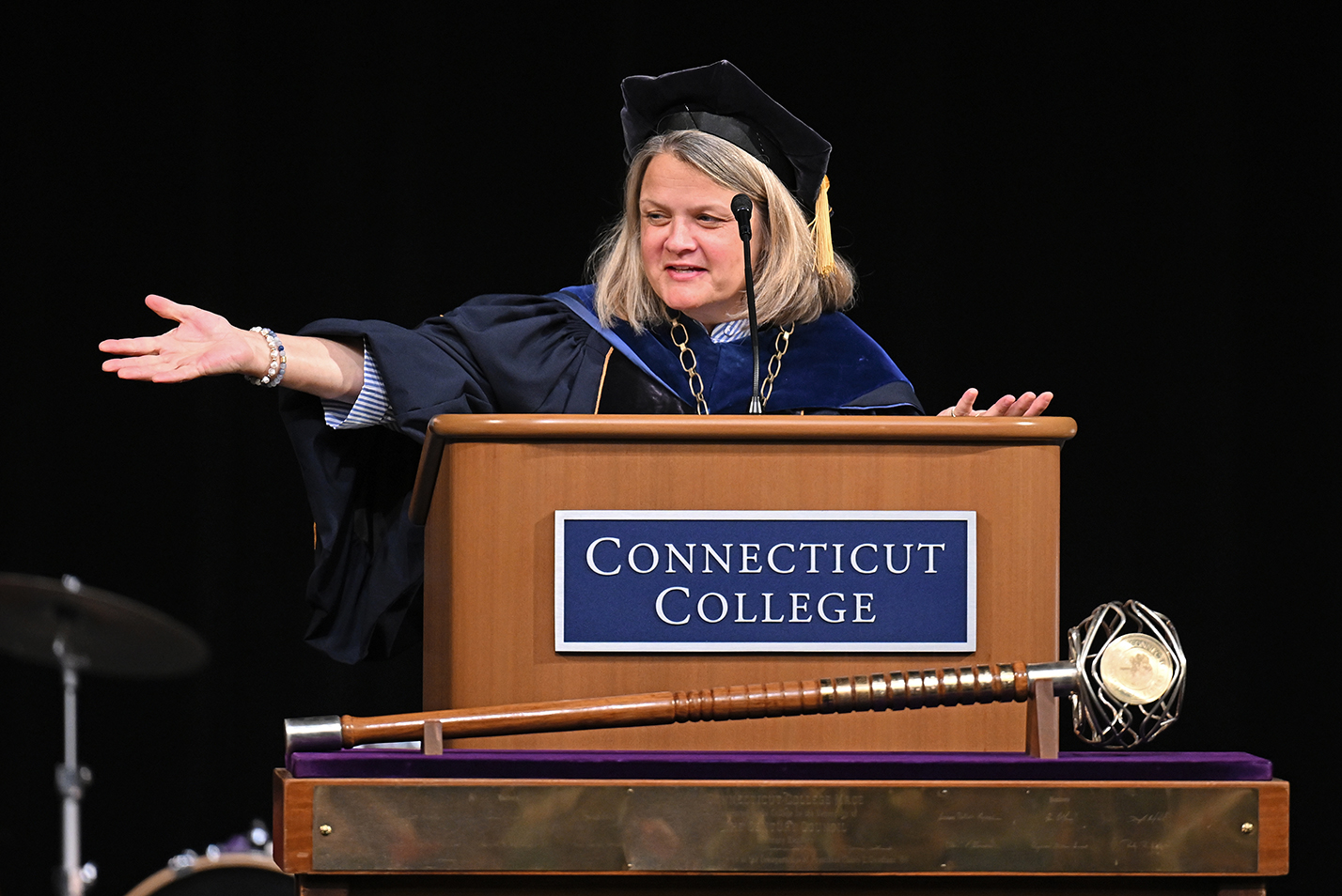 A female college president dressed in academic regalia stands behind a podium that reads Connecticut College and gestures as she speaks.