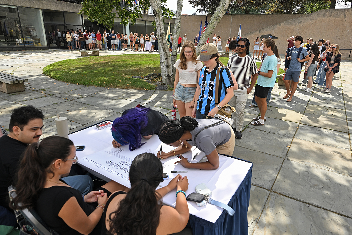 College students lean over to sign a poster-size document in an outdoor courtyard.