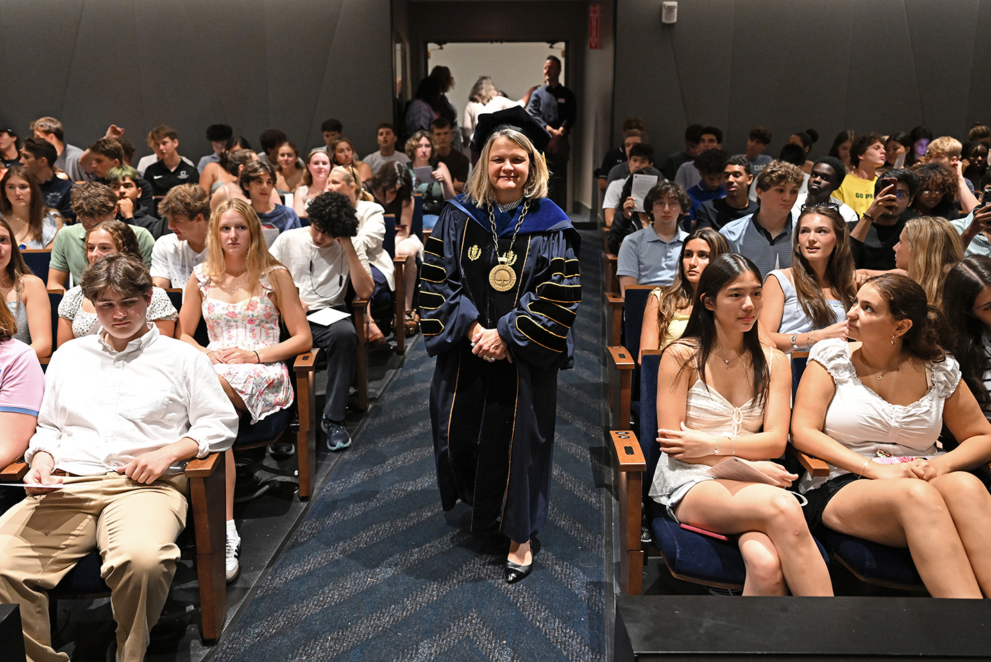 A college president in academic regalia walks down the aisle entering an auditorium between rows of seated students.