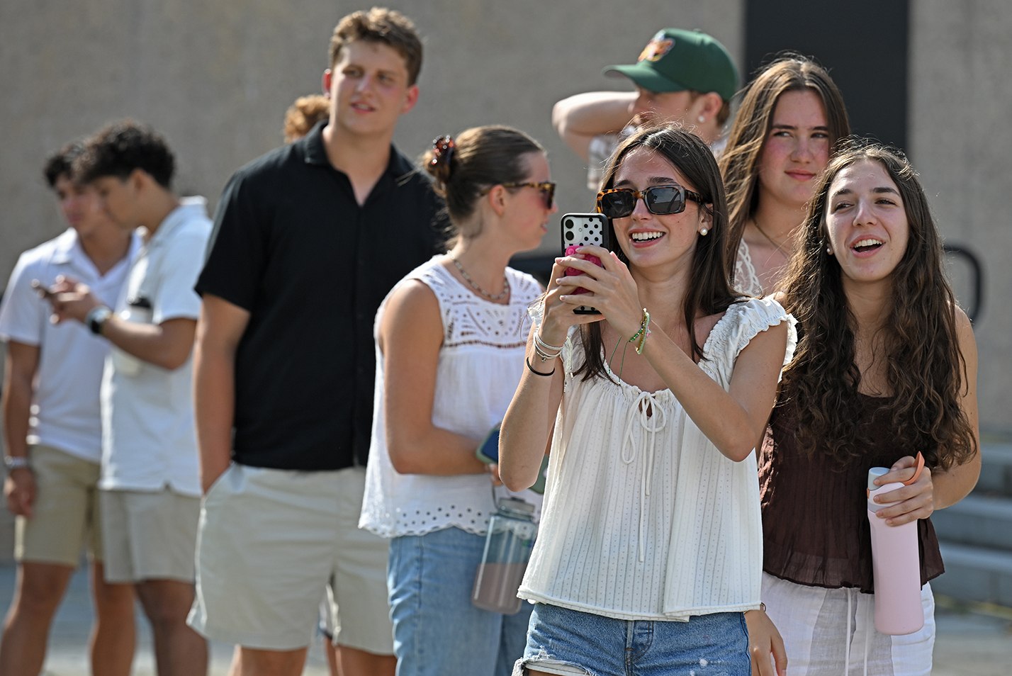 College students in casual attire smile as one takes photos with a smart phone of an activity not visible in the frame.
