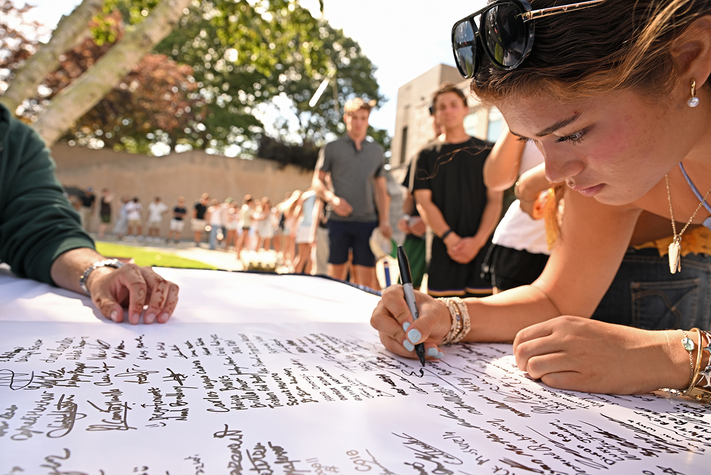 A college students leans over to sign a poster-size document in an outdoor courtyard.