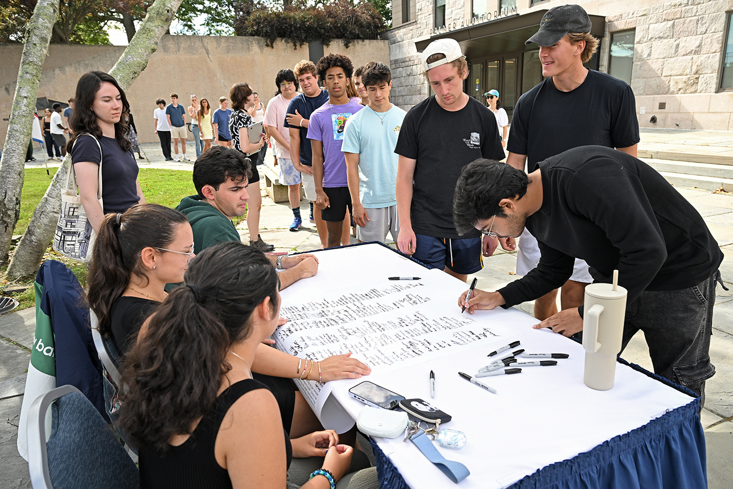 College students lean over to sign a poster-size document in an outdoor courtyard.