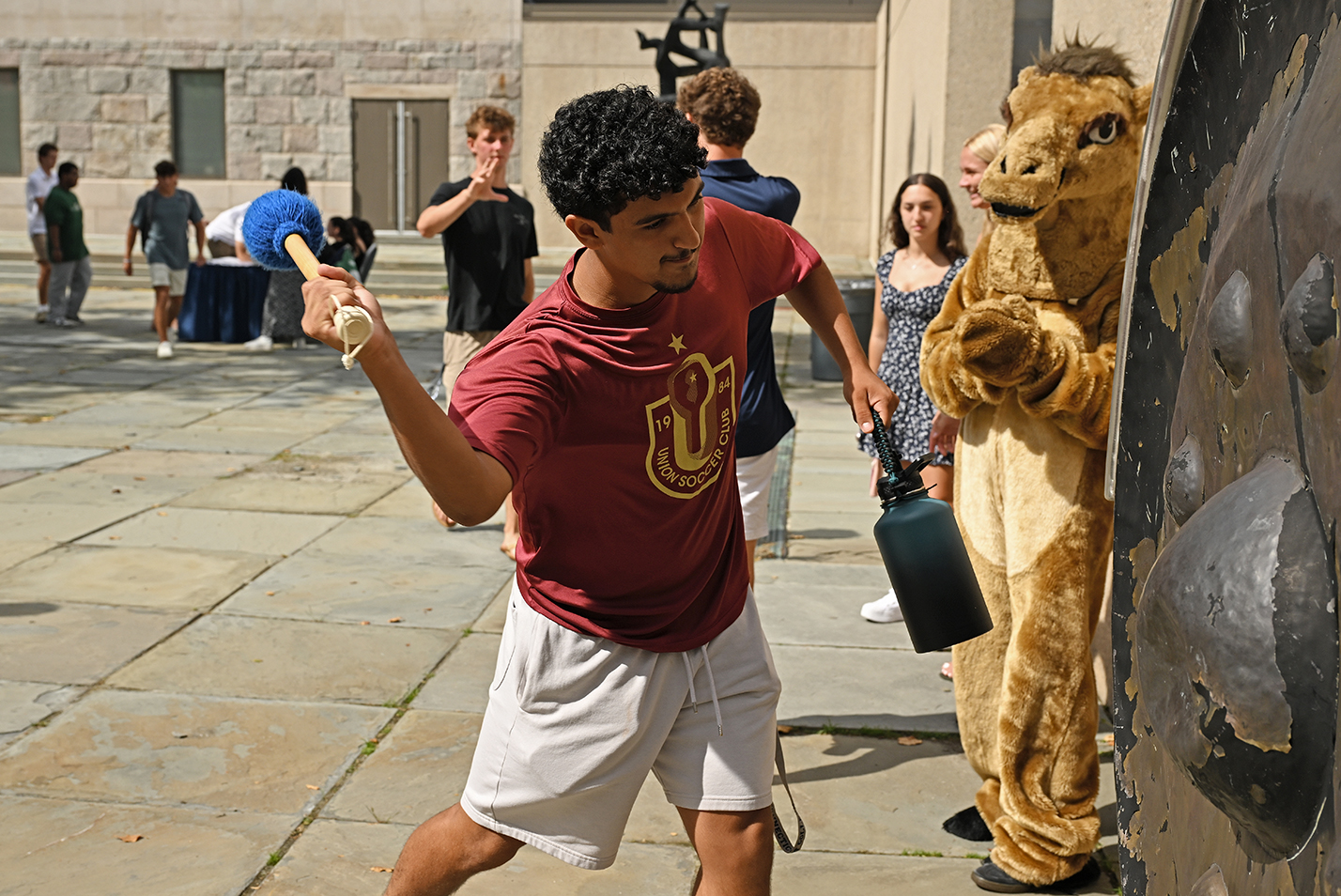 A college student in red t-shirt and khaki shorts and water flask in left hand swings a large blue padded mallet to ring a gong as fellow students, including one dressed in a camel mascot costume, watch in an outdoor courtyard.