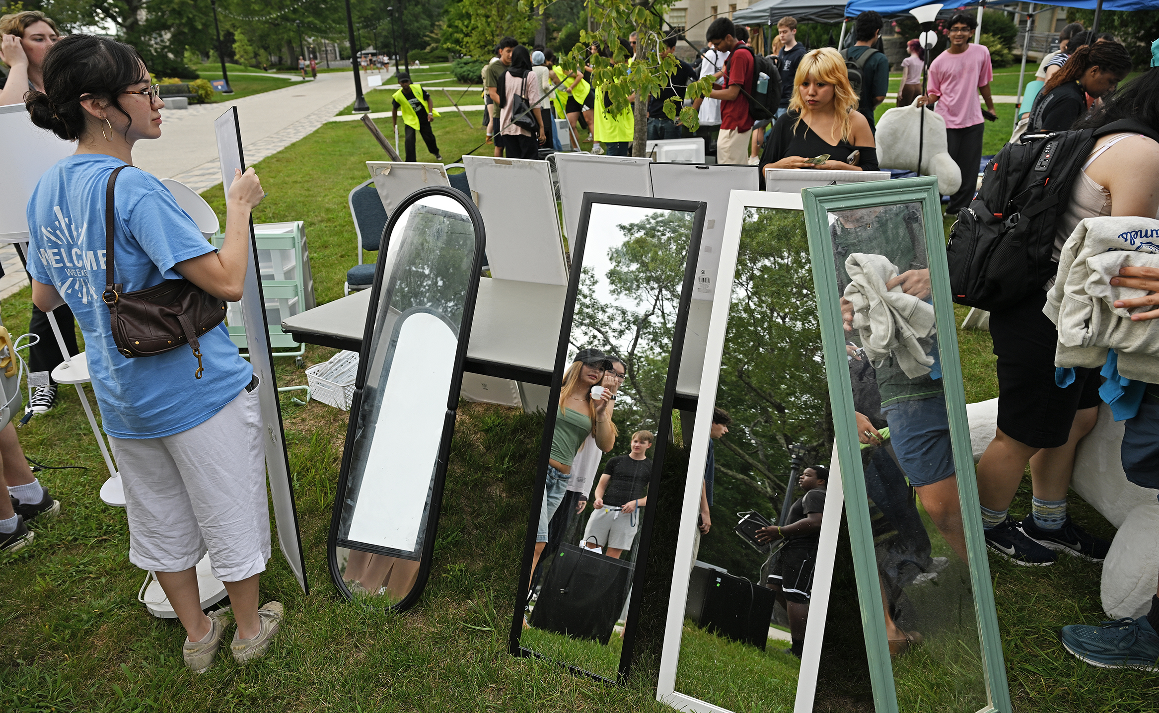 A female college student holds a mirror and watches as others are reflected in a row of tall mirrors on display during an outdoor yard sale as other students look at the various items for sale.
