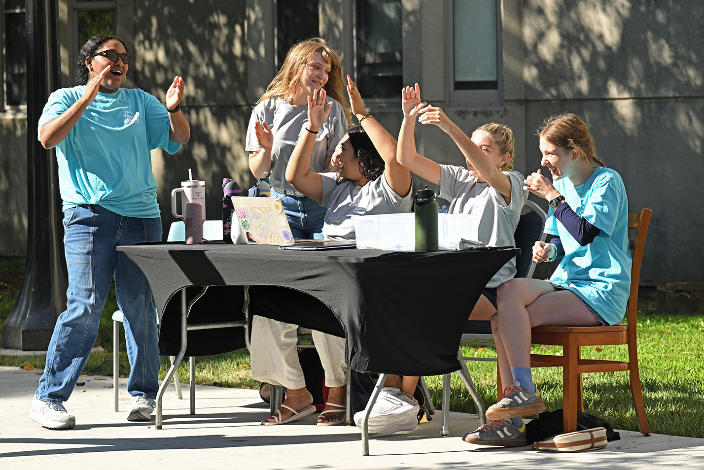 College residence life staff sits at a table and sings and waves arms as they wait to check students into their residence hall.