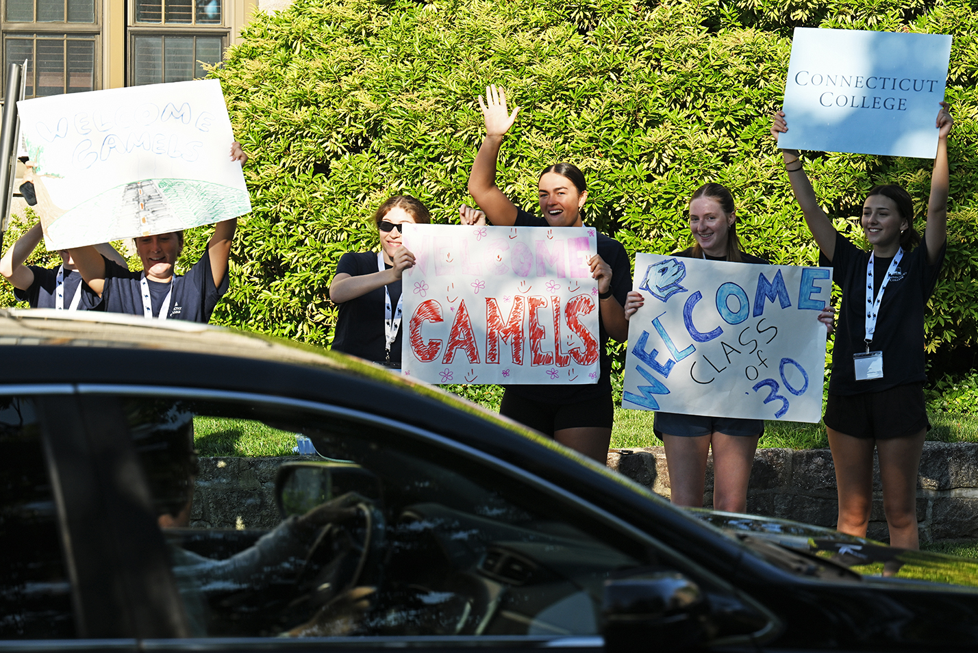 College students wave banners and smile and cheer at a car pulling onto campus for move-in day.