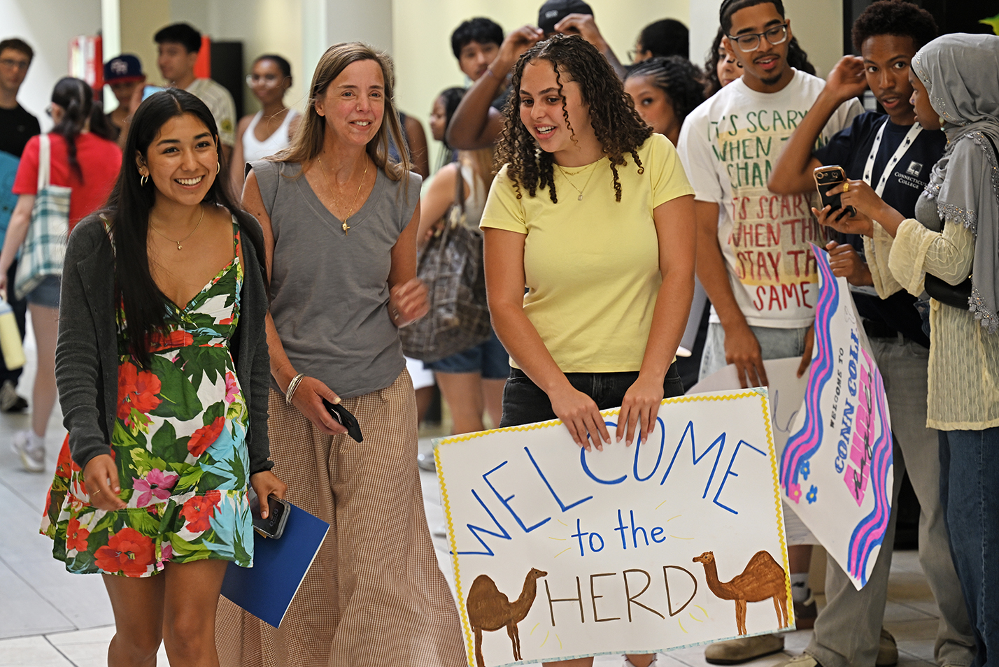 A college student holding a hand-made welcome sign walks with a new student through a crowded corridor.