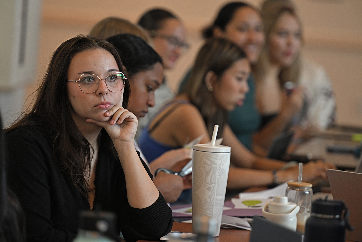 A female college student in glasses rests her chin in her hand as she and a group sitting at a long table listen to a speaker.