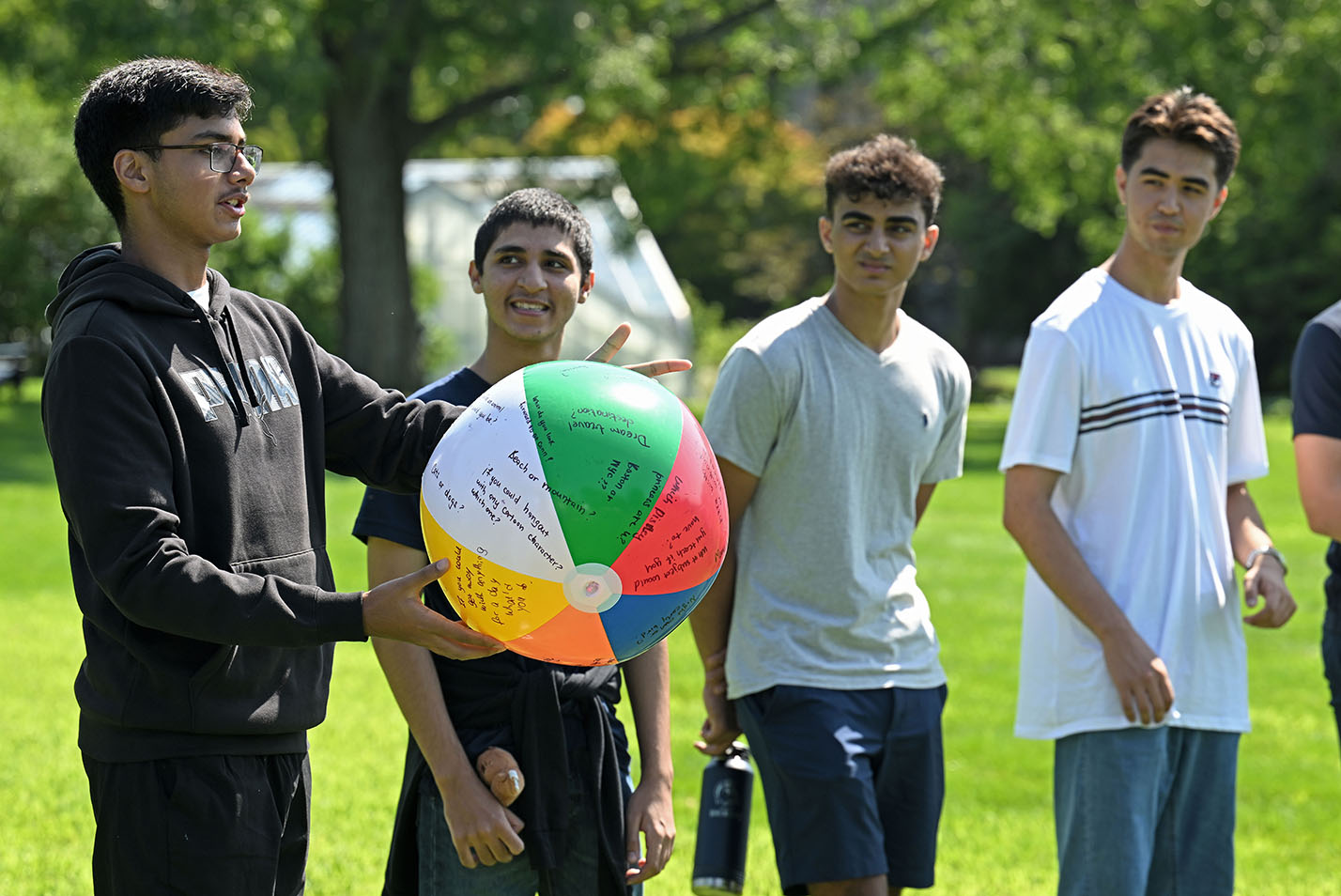 A male college student holds a multi-color beach ball with questions written on it and speaks to a group of students outside on a college green.