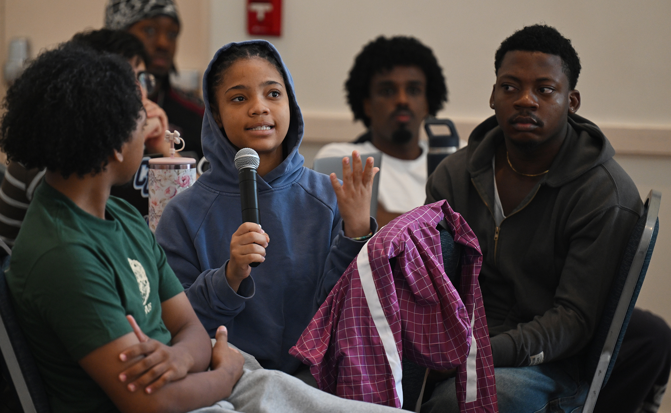 A college student in a blue hooded sweatshirt hold a microphone and gestures as she asks a question.