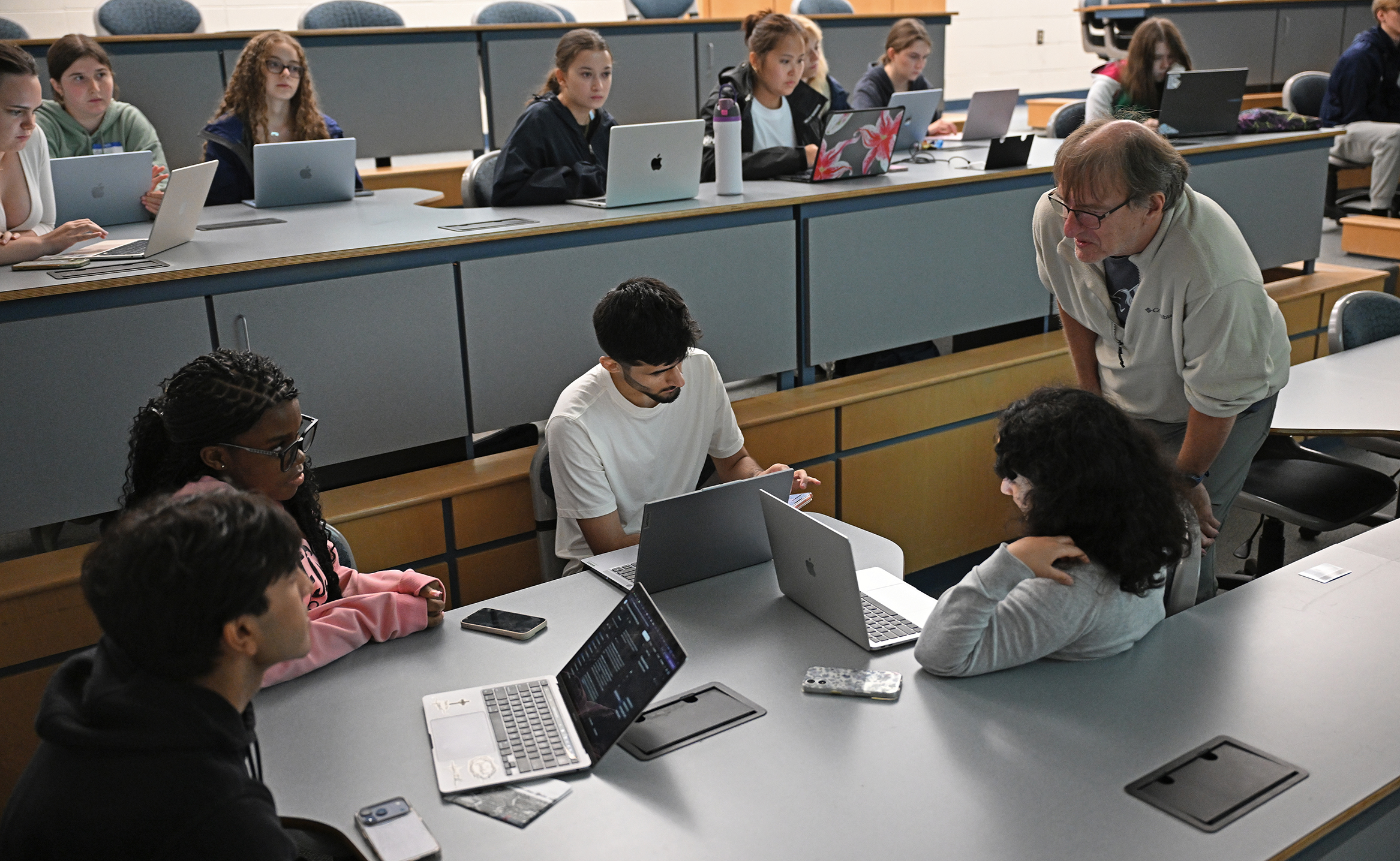 A college professor leans over to speak to a group of students gathered around their laptop computers in a lecture hall.