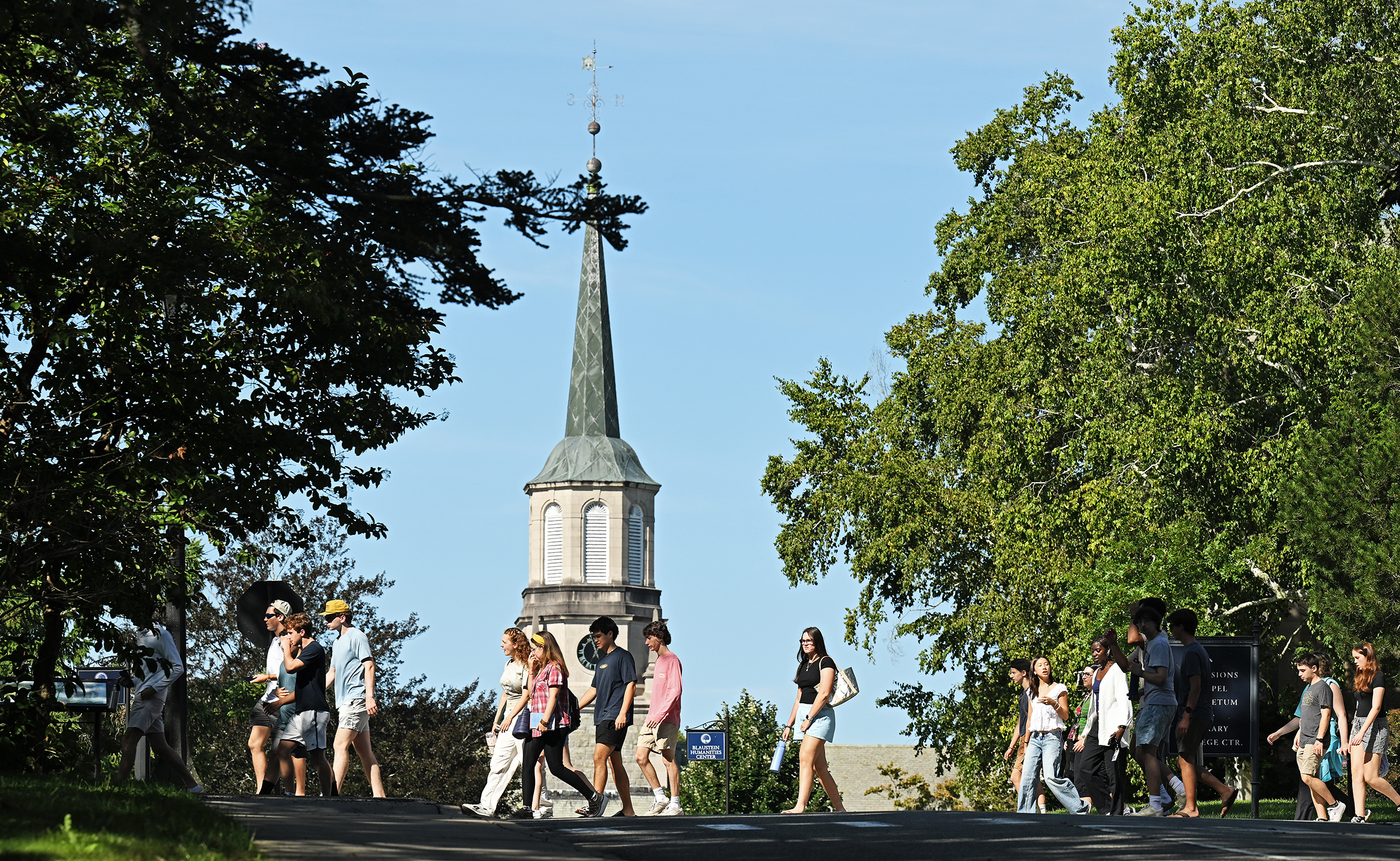 College students cross a campus street with a chapel steeple visible in the distance.
