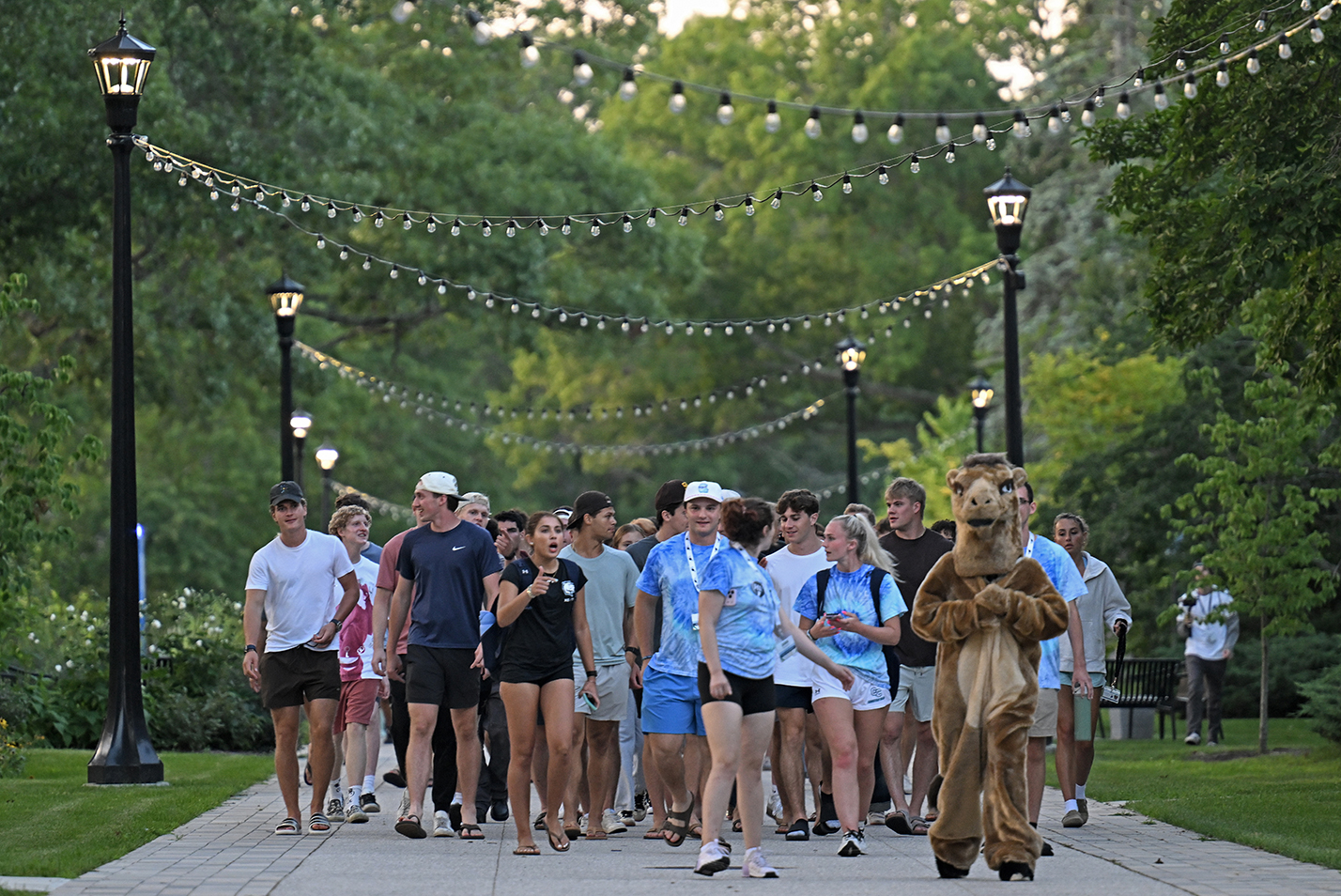 A crowd of students walk down a pedestrian promenade led by a student dressed in a camel mascot costume.