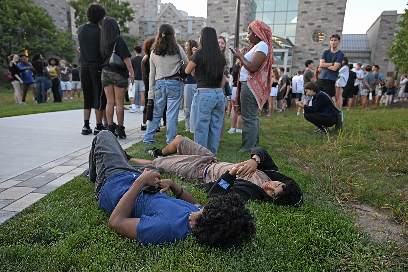 two college students lay down in the grass as their classmates gather on the lawn outside a dorm building at dusk.