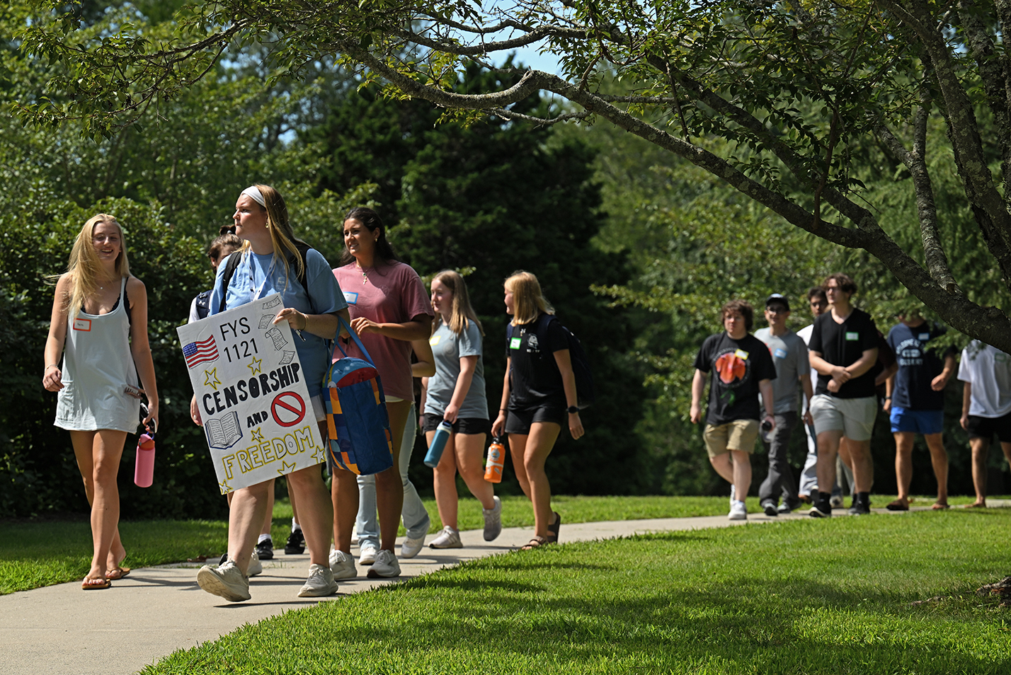 College students, one carrying a hand-made poster, walk along a pathway on campus.