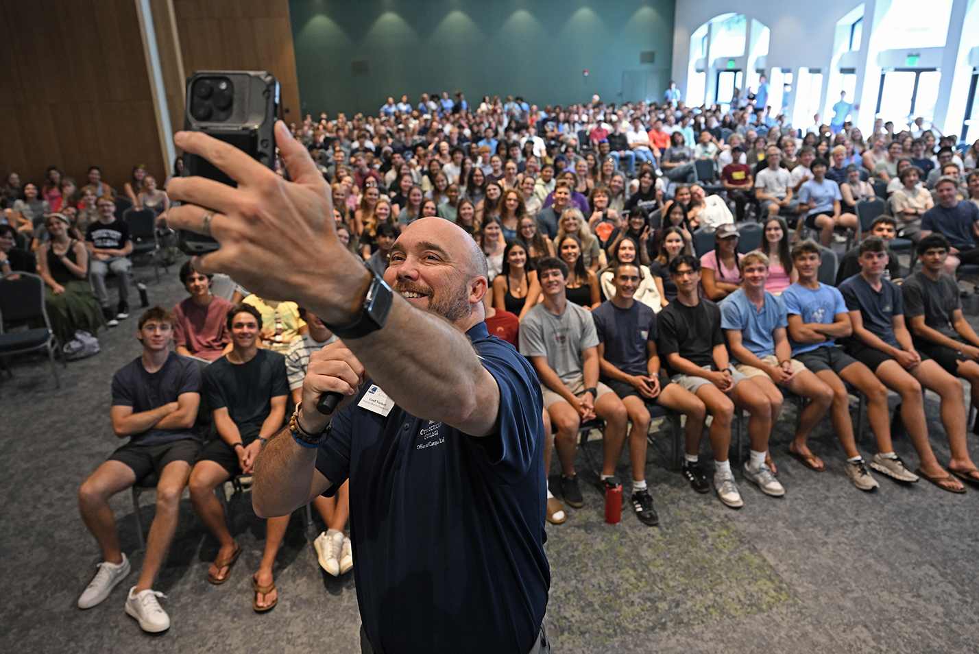 A college dean takes a selfie photo with an auditorium full of first year students.