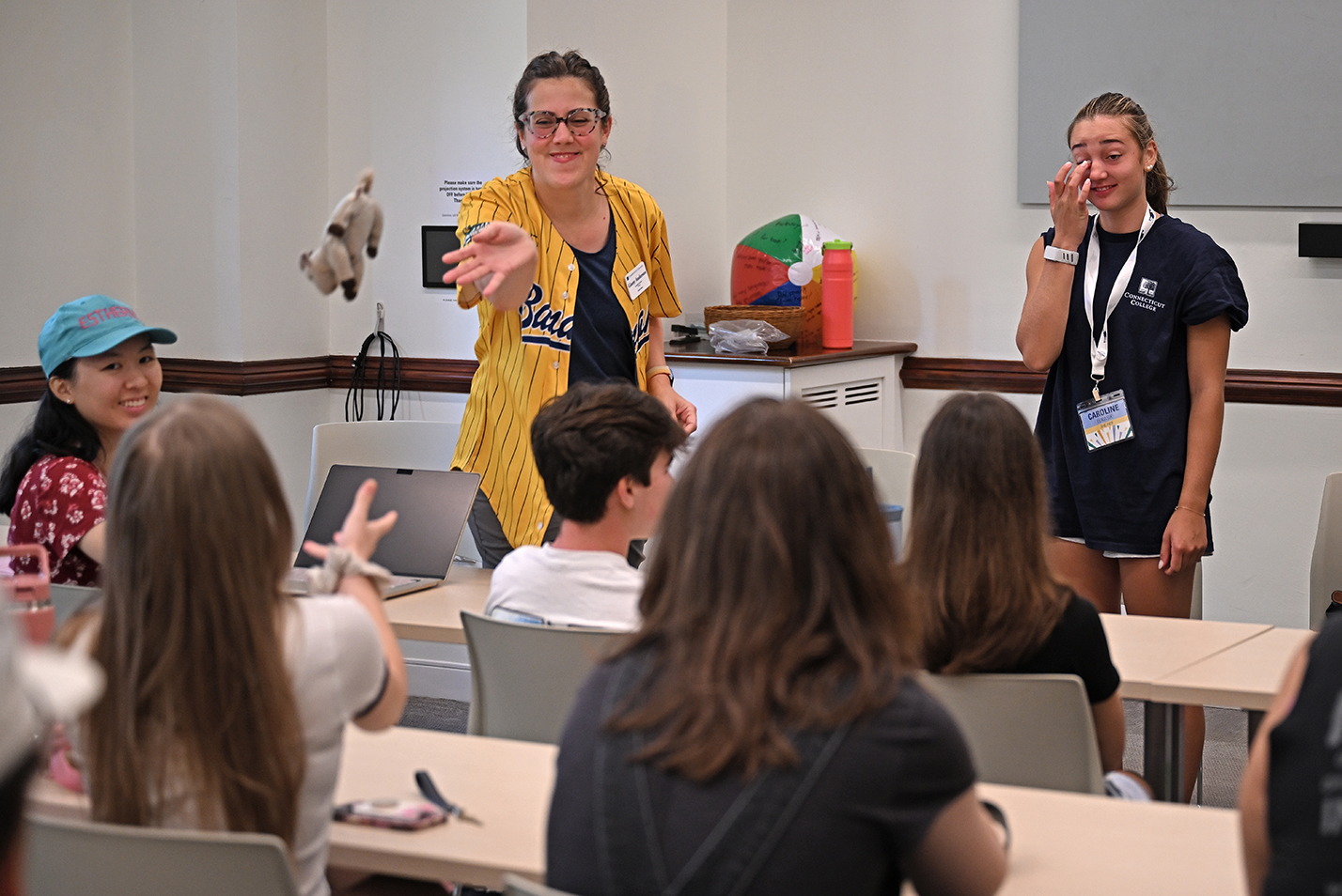 A college professor in a yellow baseball jersey tosses a small stuffed animal to a seated student during an orientation session.